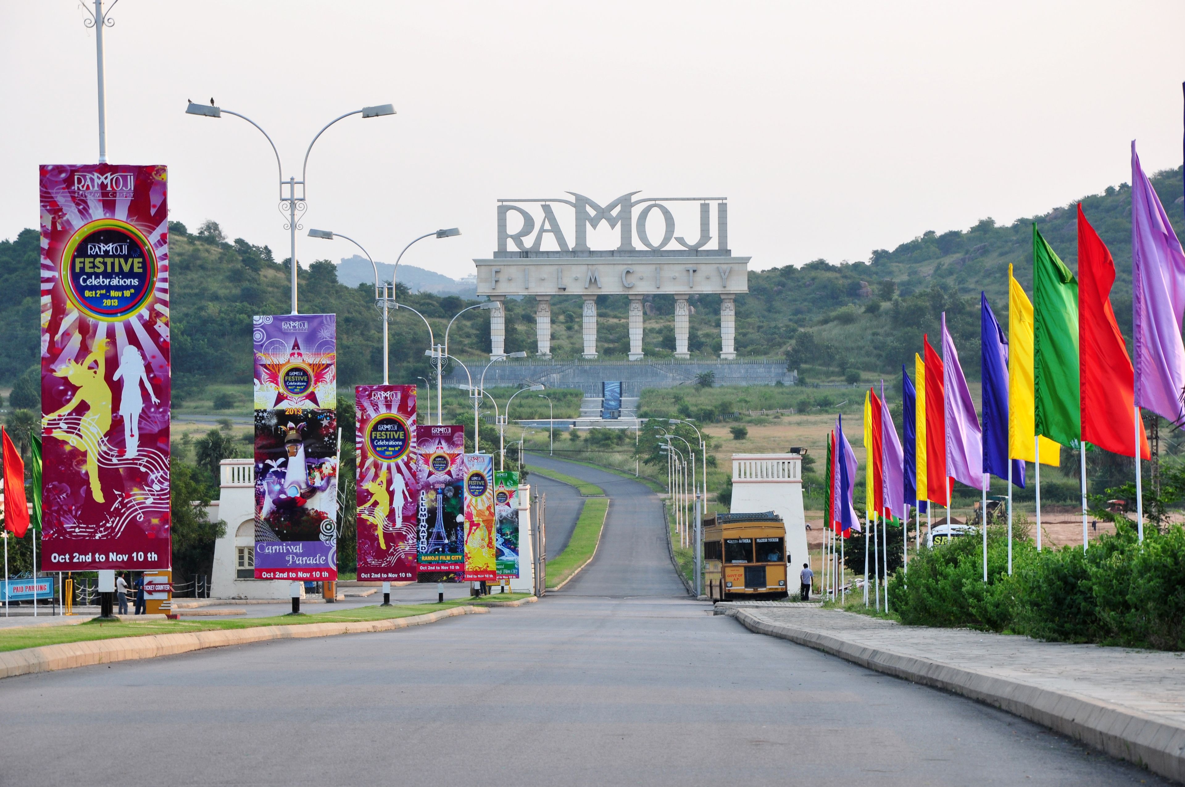 A road leading to a large building with the name 'RAMOJI' on it, surrounded by colorful flags and banners advertising a festival.