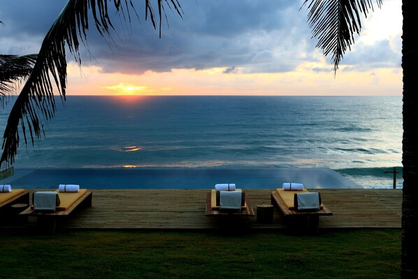 Une terrasse en bois avec des chaises longues et une vue sur l’océan au coucher du soleil.