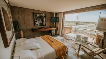 Quarto com cama grande, televisão, janela com vista para a praia e uma cadeira de madeira.