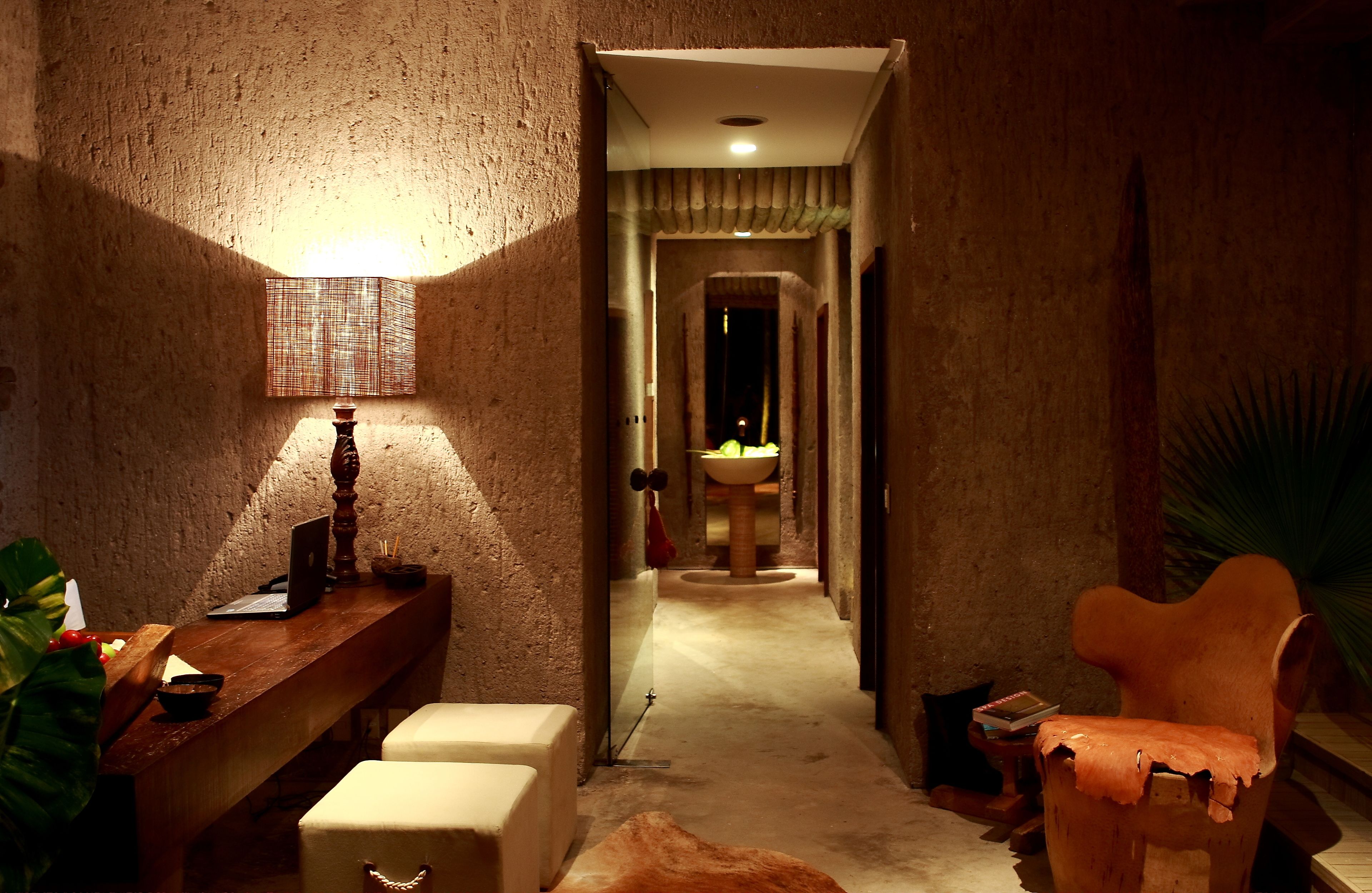 A narrow hallway with a wooden bench, a desk with a laptop, a lamp, and a chair. A sink is visible in the distance.