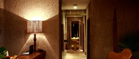 A narrow hallway with a wooden bench, a desk with a laptop, a lamp, and a chair. A sink is visible in the distance.