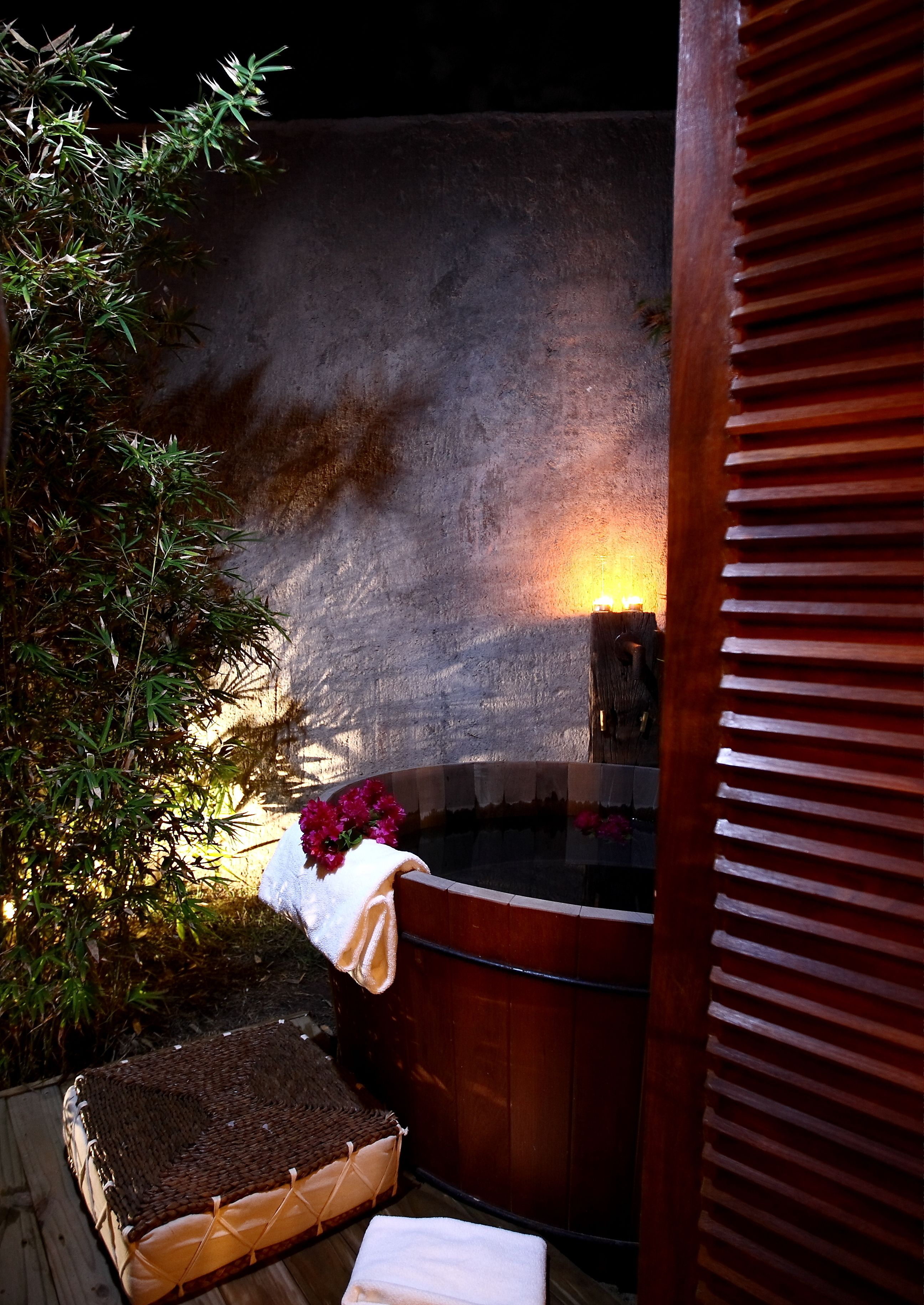 Un jacuzzi con un banco de madera y una toalla, rodeado de follaje y un muro de piedra.