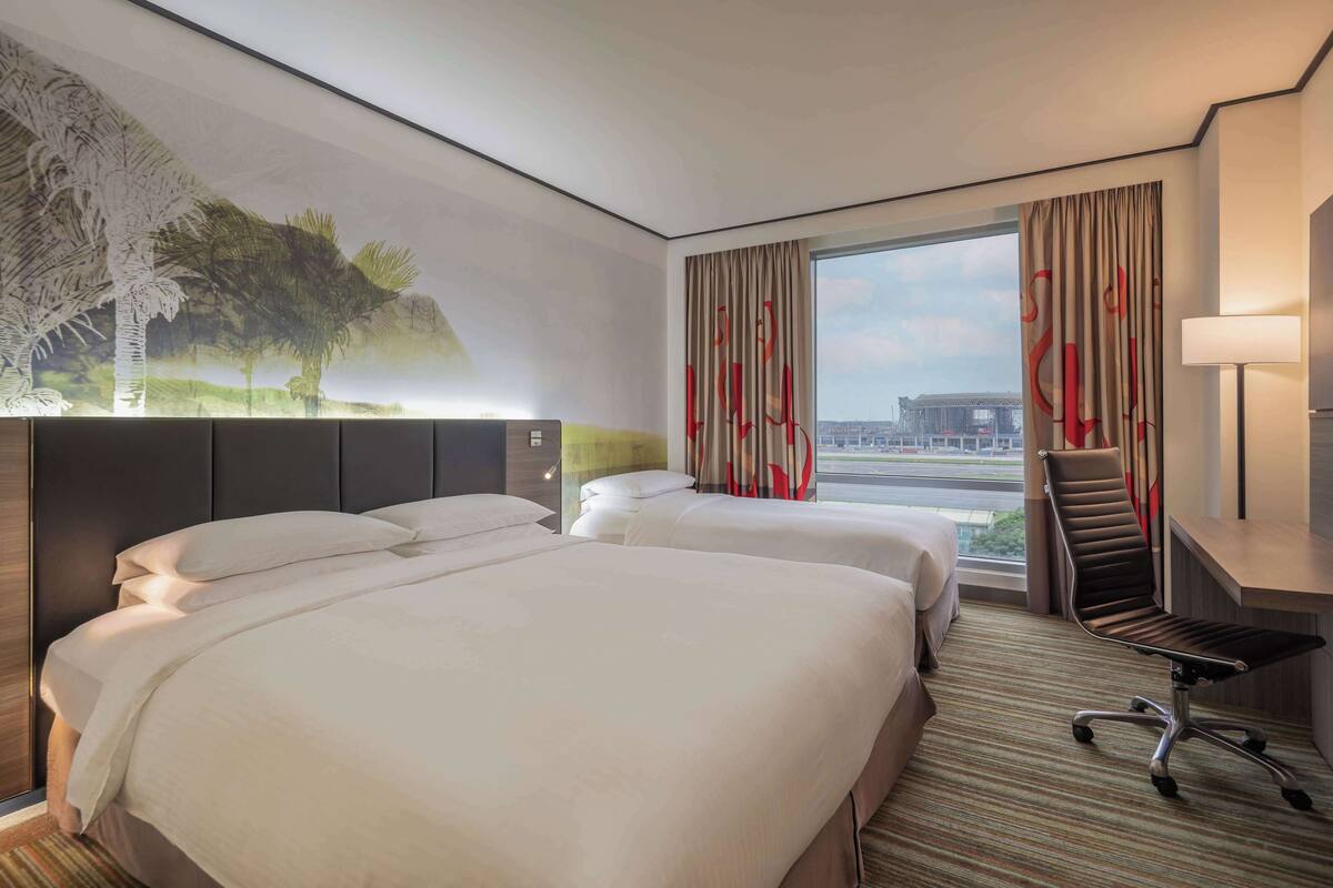 A hotel room with two beds, a desk, a chair, and a large window with curtains.