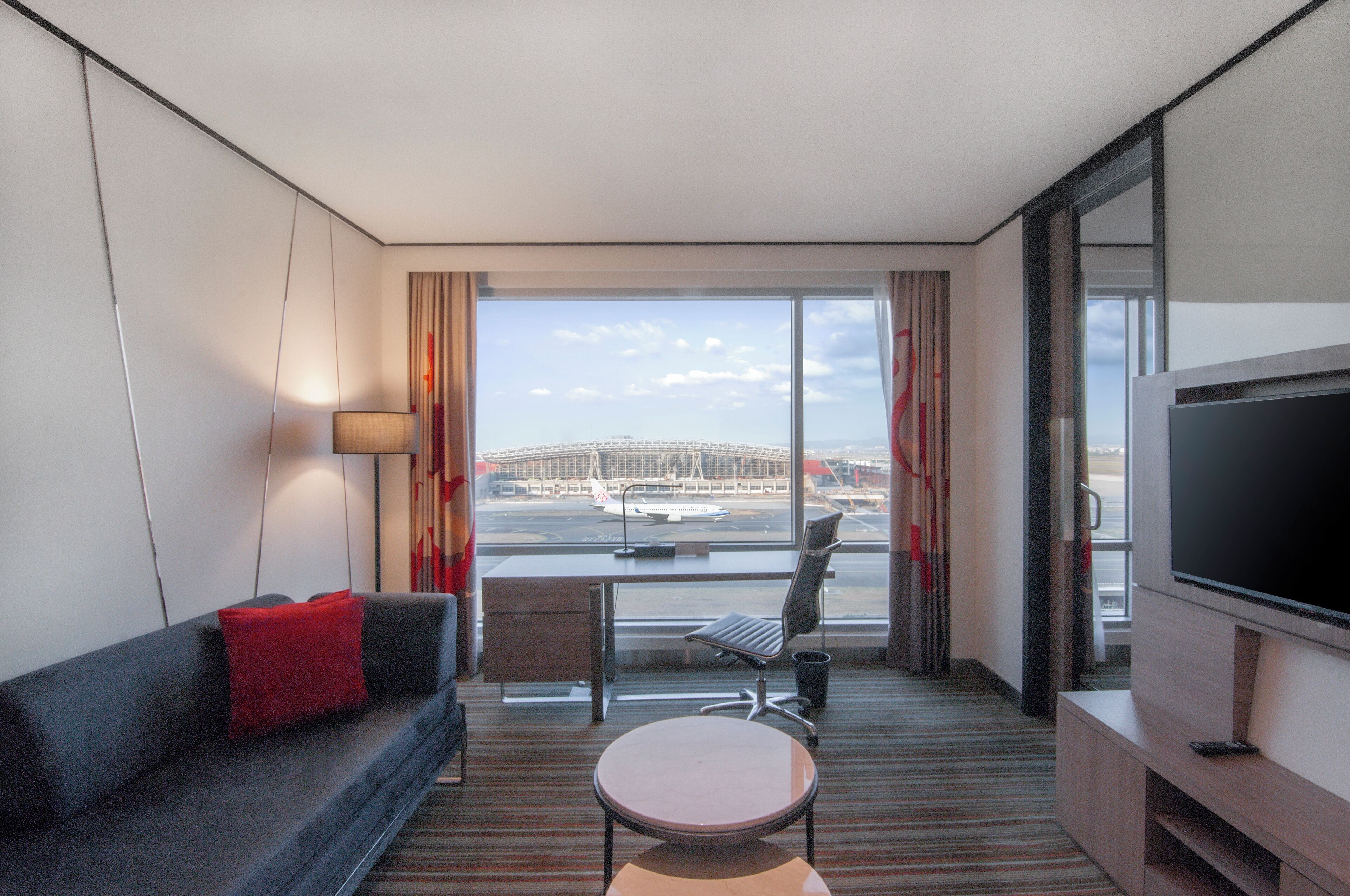 Habitación de hotel con sofá, una mesita, televisor y vistas al aeropuerto.