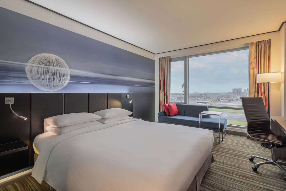A hotel room with a large bed, a desk, a chair, a sofa, and a view of the cityscape.