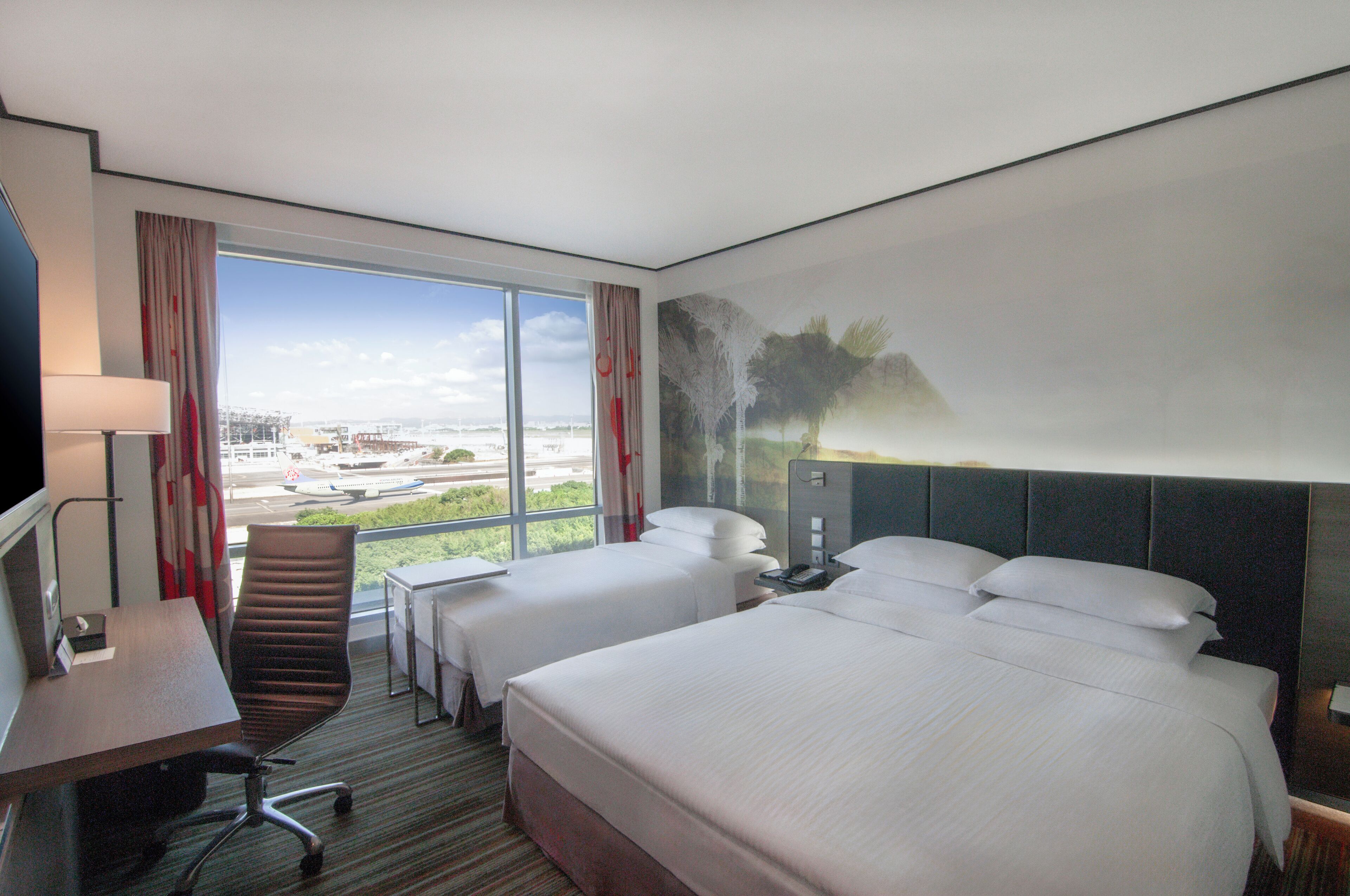 A hotel room with two beds, a desk, a chair, and a large window offering a city view.