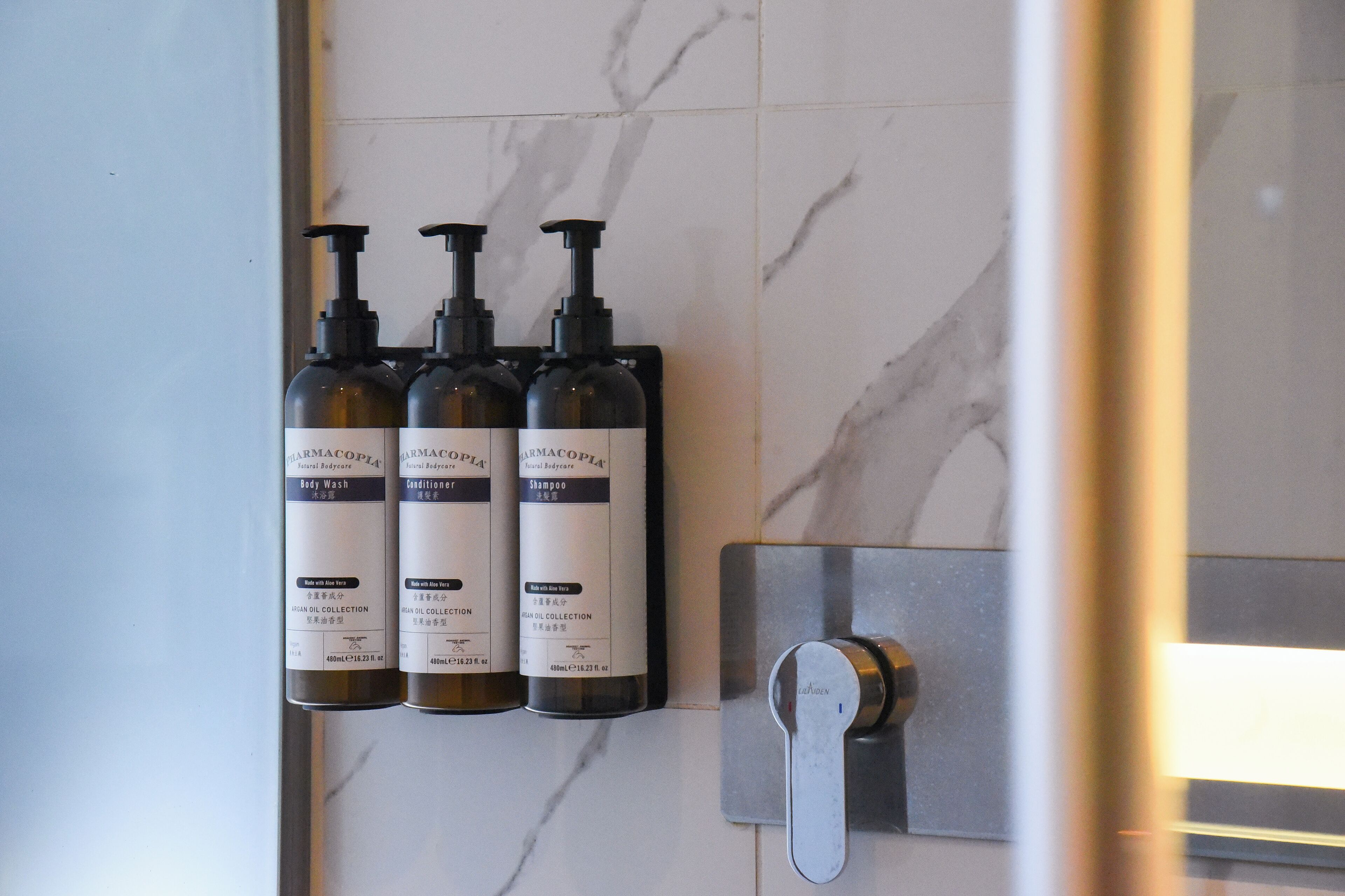 A bathroom with a marble wall and three bottles of body wash, conditioner, and shampoo.