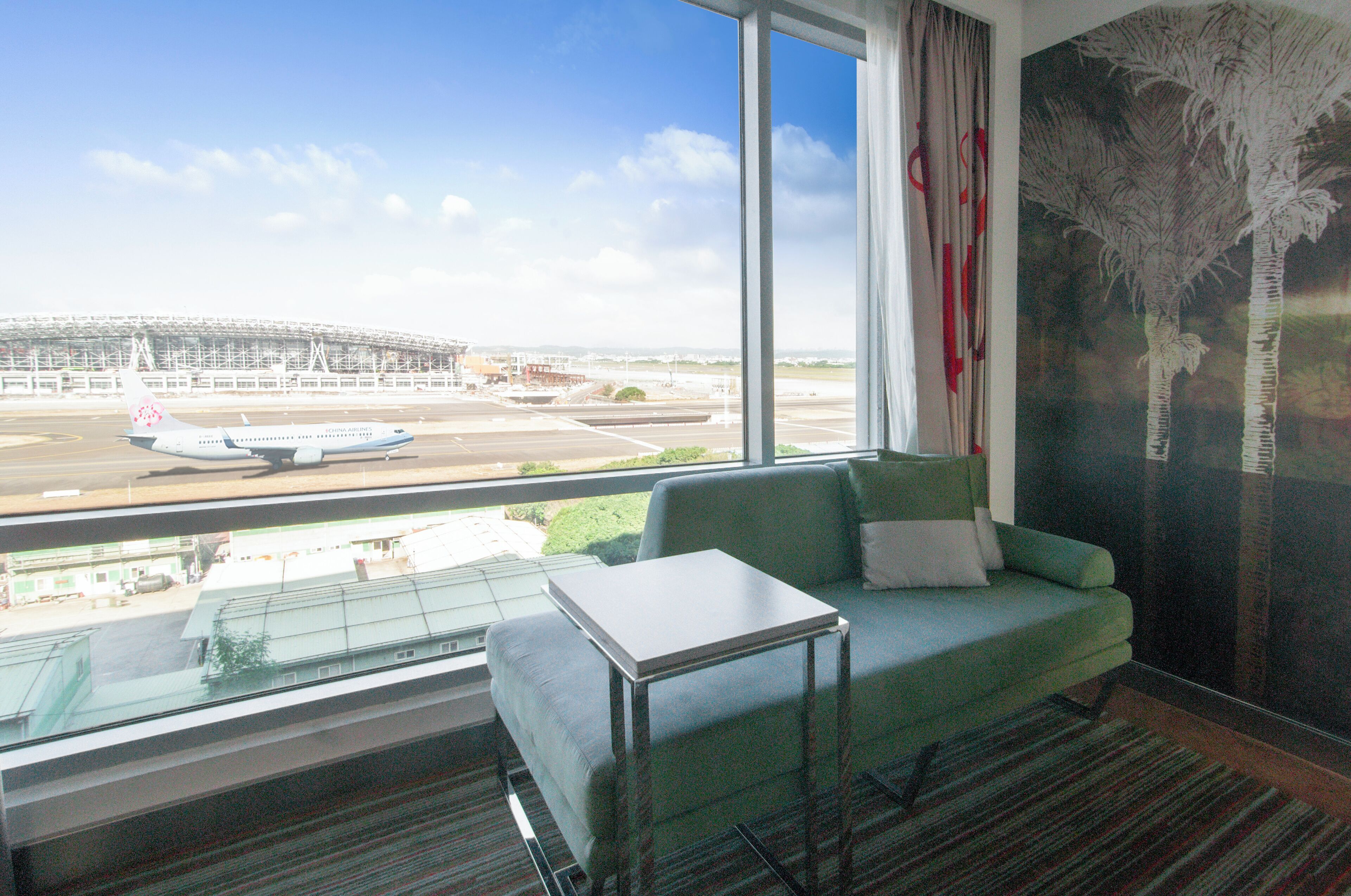 Een kamer met een groot raam met uitzicht op een luchthaven, een groene bank en een glazen bijzettafeltje.