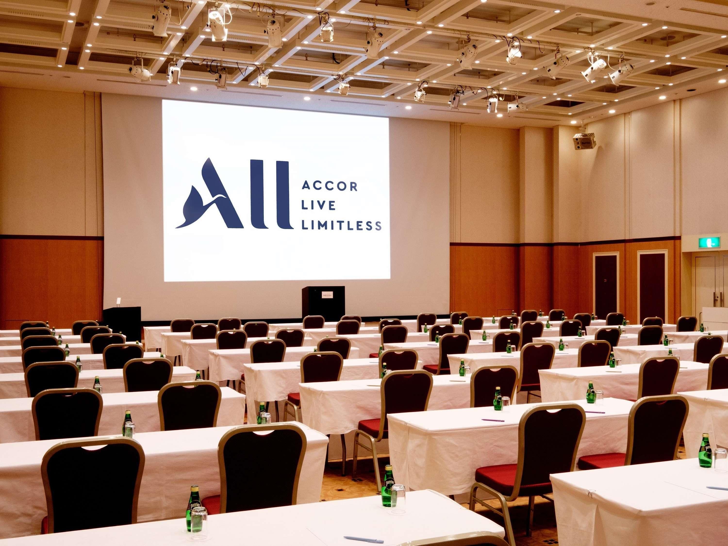 A conference room with rows of tables and chairs, a large screen displaying 'All ACCOR LIVE LIMITLESS', and a speaker at the front.
