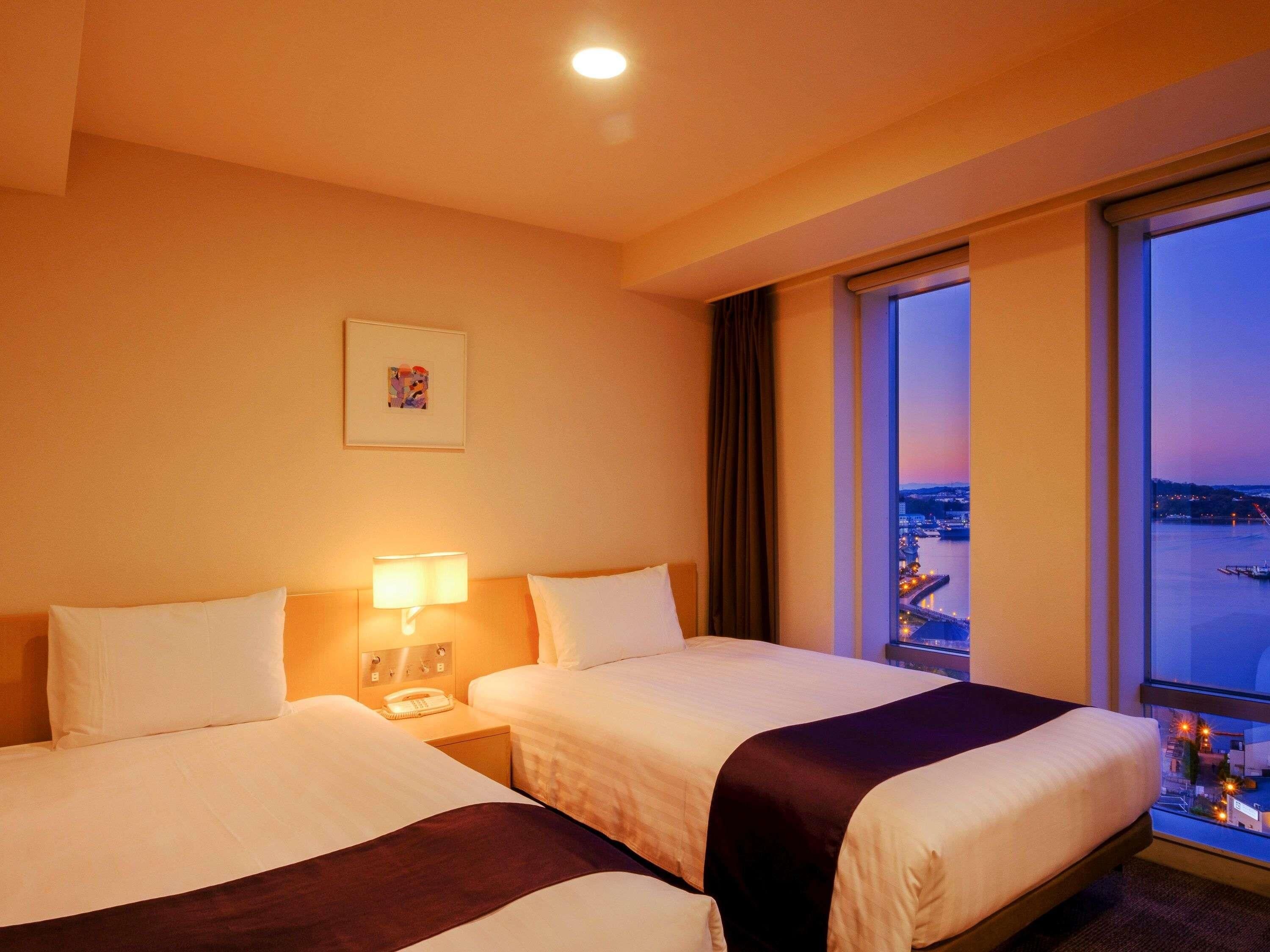 A hotel room with two beds, a nightstand with a lamp, a framed picture on the wall, and a large window offering a city view.