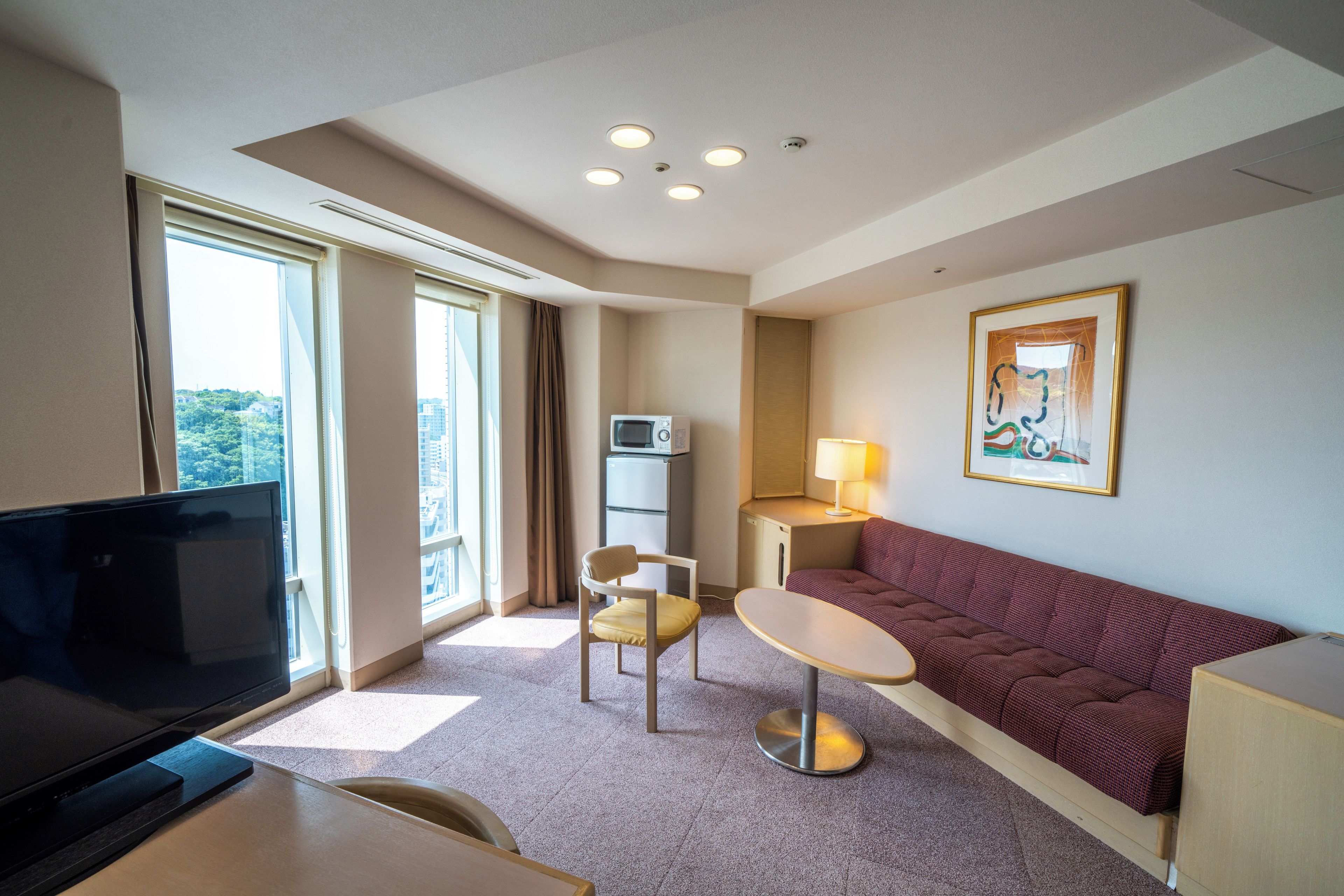 Quarto de hotel com televisão, sofá, mesa pequena, cadeira, luminária e um quadro na parede.