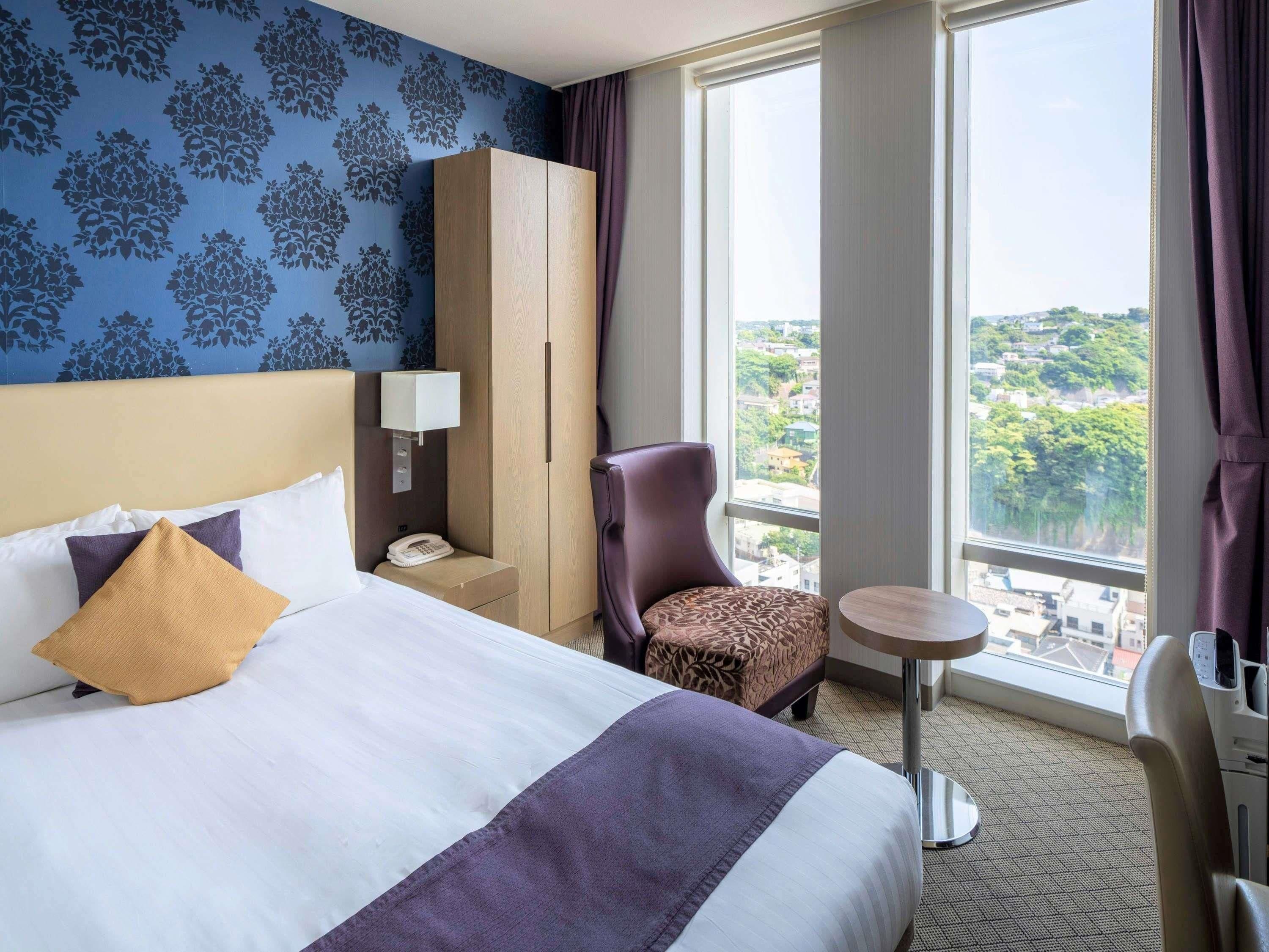 A hotel room with a large bed, a wardrobe, a desk with a lamp, a chair, and a view of the city.
