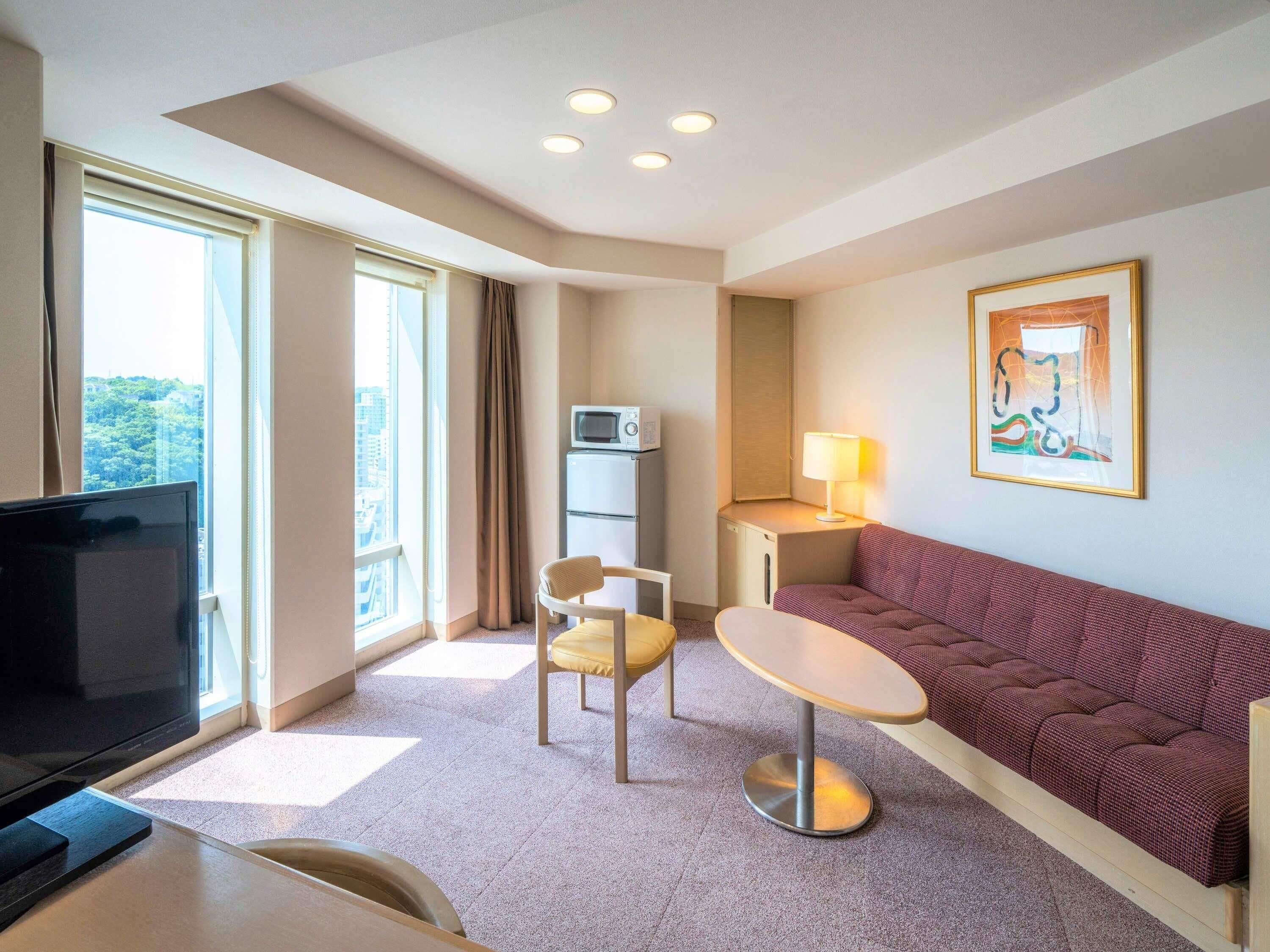 A hotel room with a TV, a sofa, a small table, a chair, a lamp, and a painting on the wall.