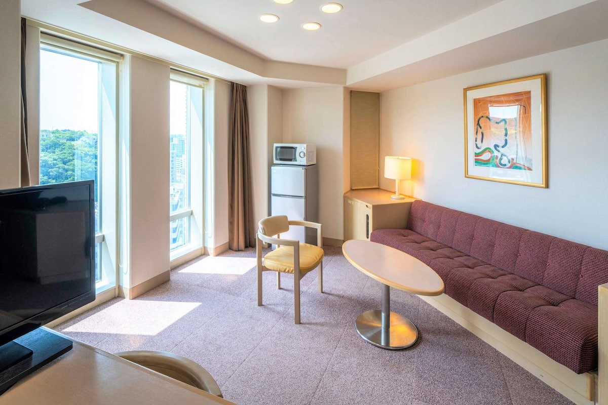 A hotel room with a TV, a sofa, a small table, a chair, a lamp, and a painting on the wall.