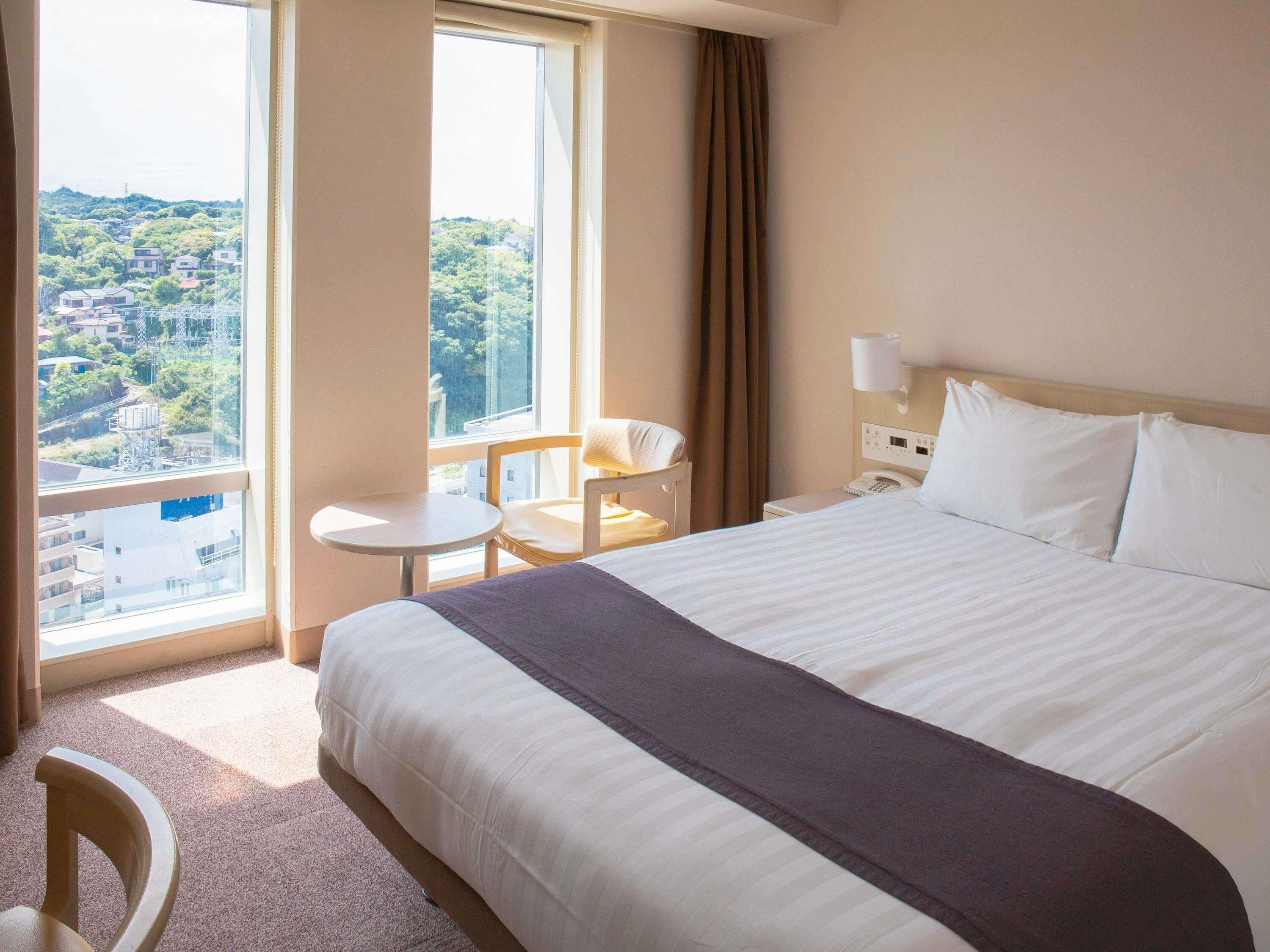 Habitación de hotel con una cama grande, una mesita, una silla, una mesita de noche y una ventana con vista a un paisaje urbano.