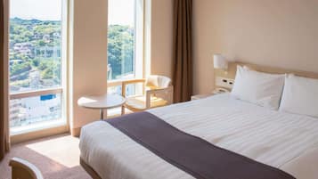 Een hotelkamer met een groot bed, een klein tafeltje, een stoel, een nachtkastje en een raam met uitzicht op een stadsbeeld.