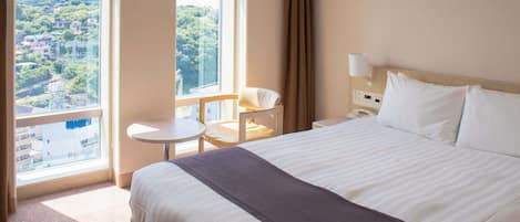A hotel room with a large bed, a small table, a chair, a nightstand, and a window with a view of a cityscape.