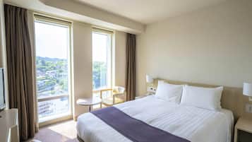 A hotel room with a large bed, a desk, a chair, and a window offering a city view.