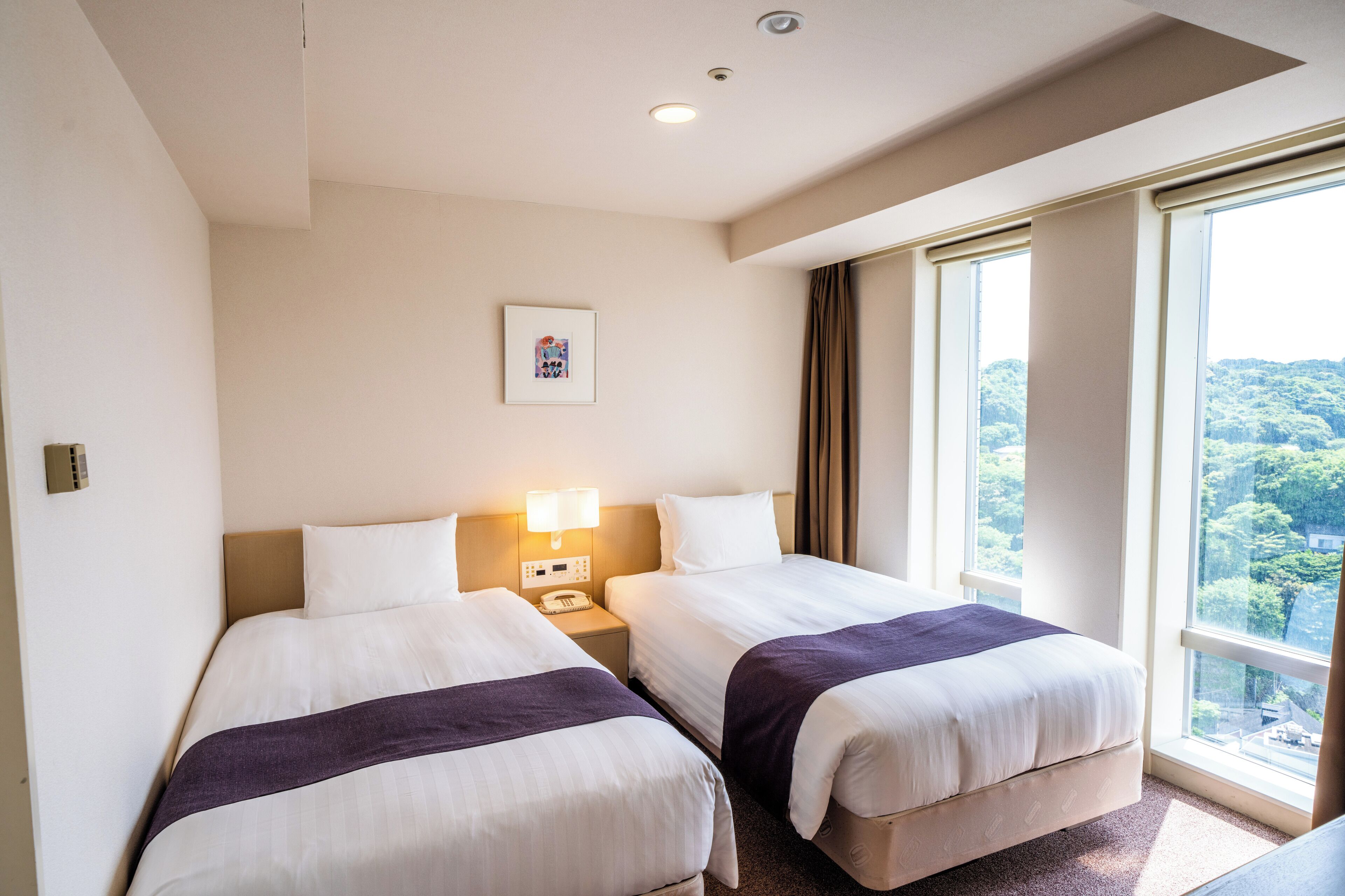 Habitación de hotel con dos camas, una mesita de noche con lámpara, un cuadro enmarcado en la pared y un amplio ventanal con vistas al exterior.
