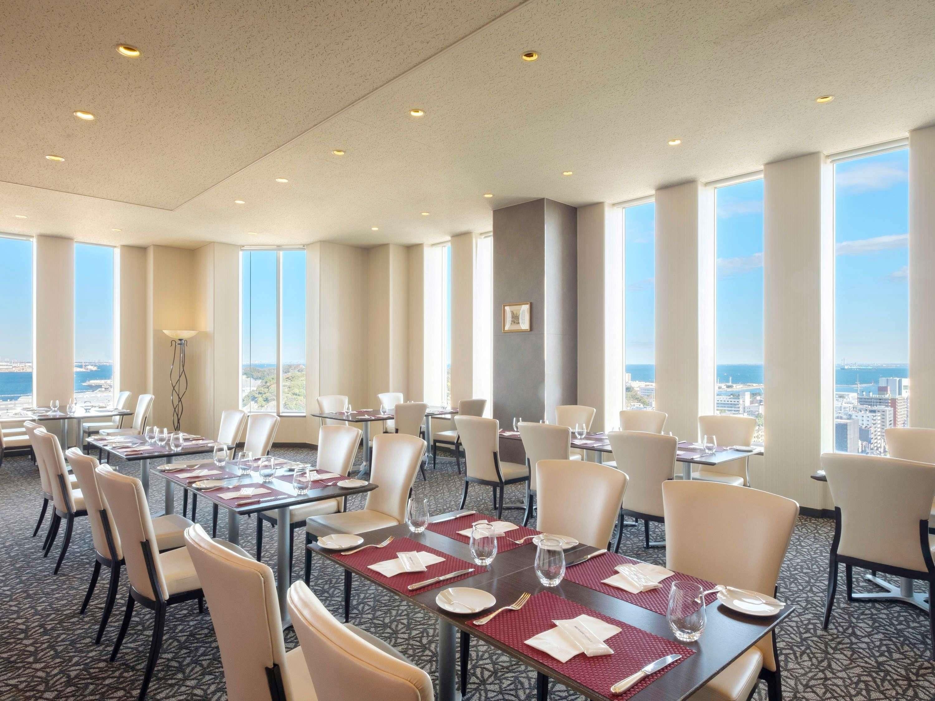 A hotel dining area with multiple tables and chairs, large windows offering a city view, and a patterned carpet.