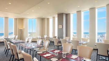 A hotel dining area with multiple tables and chairs, large windows offering a city view, and a patterned carpet.