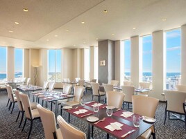 A hotel dining area with multiple tables and chairs, large windows offering a city view, and a patterned carpet.