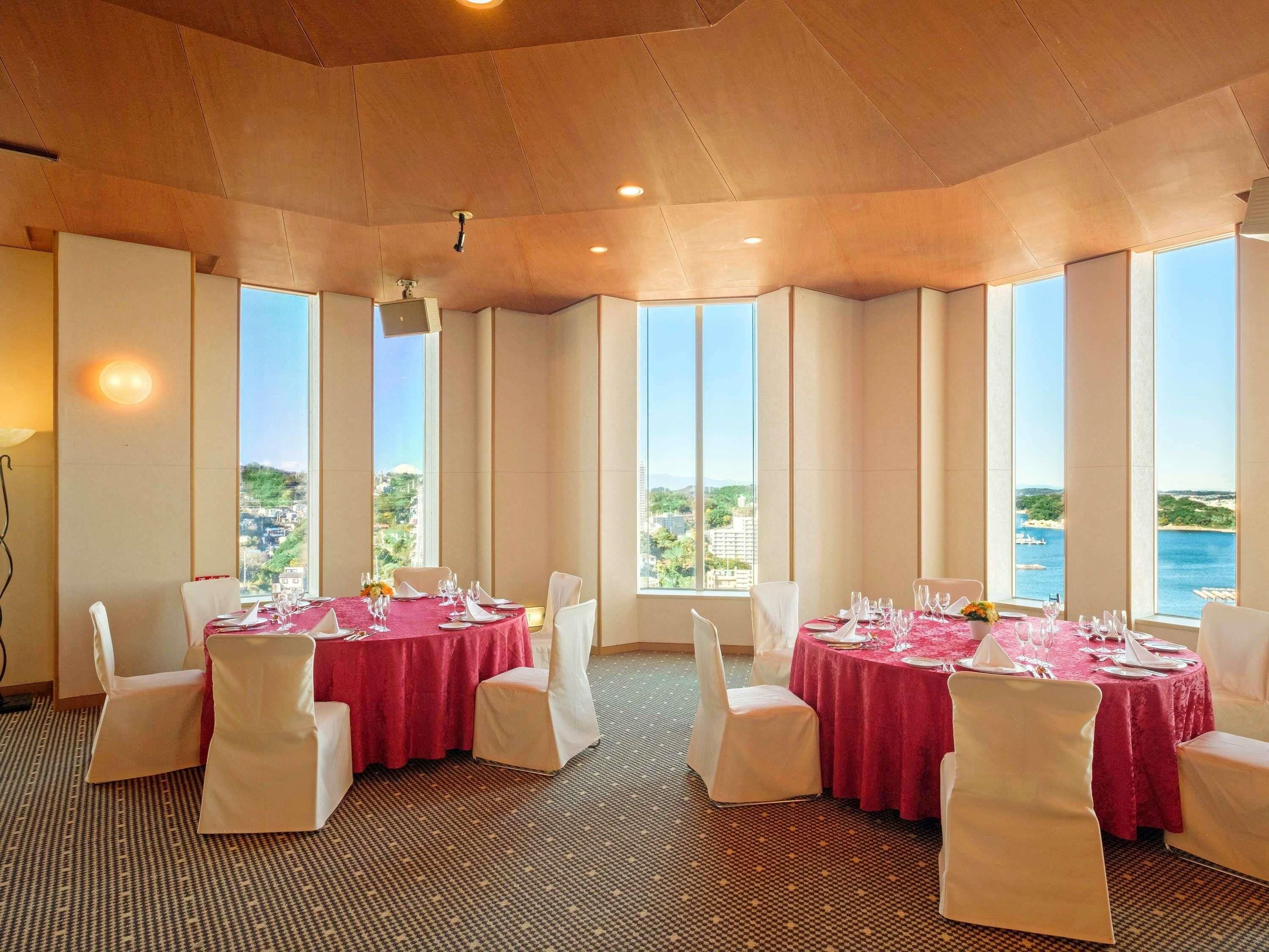 A dining area with tables set for four, large windows with a view of the sea, and a patterned carpet.