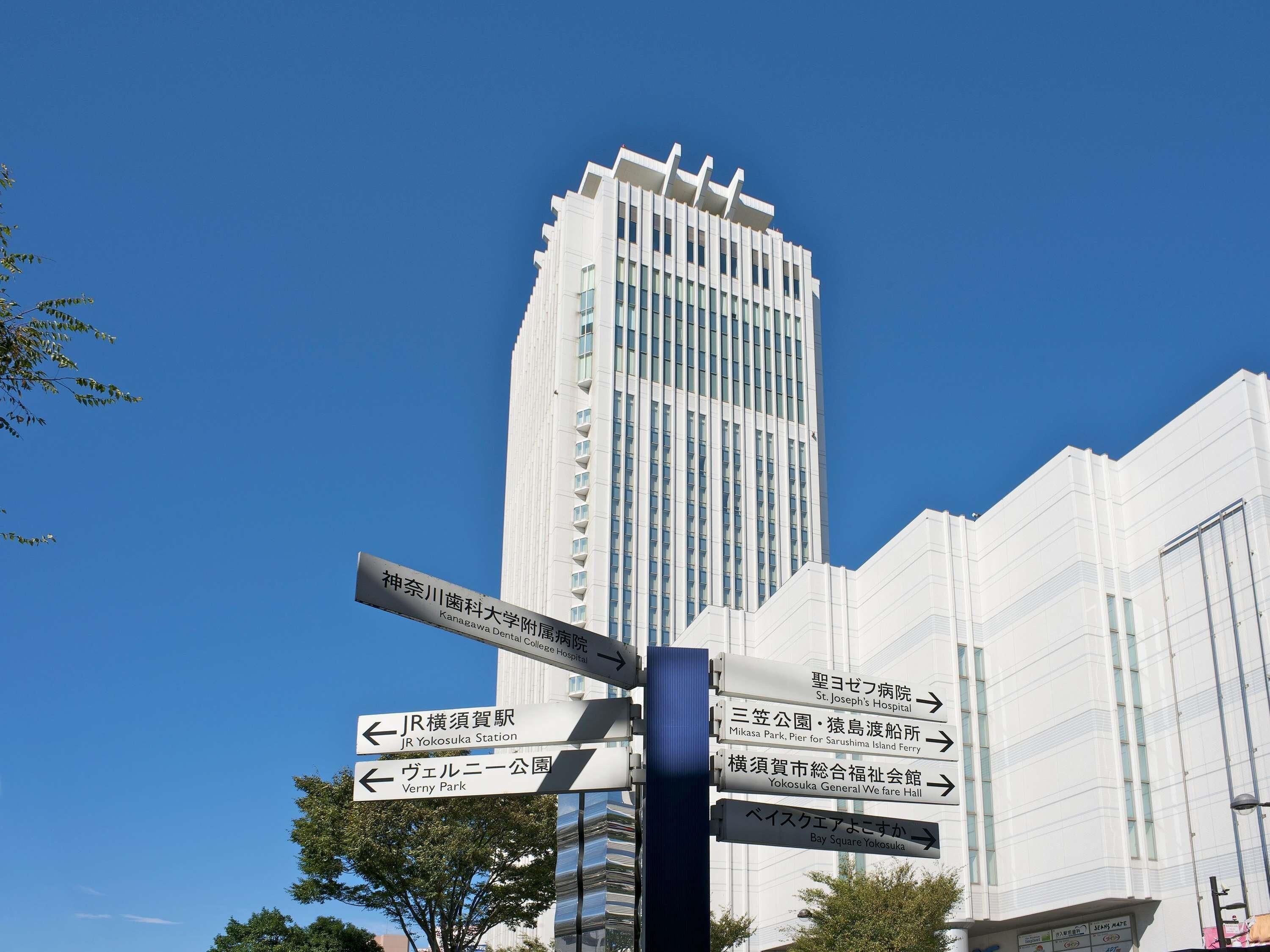 A directional signpost with multiple arrows pointing towards various destinations including JR Yokosuka Station, Verney Park, St. Joseph's Hospital, Mikasa Park, Pier for Sarushima Island Ferry, Yokosuka General We fare Hall, and Bay Square Yokosuka.