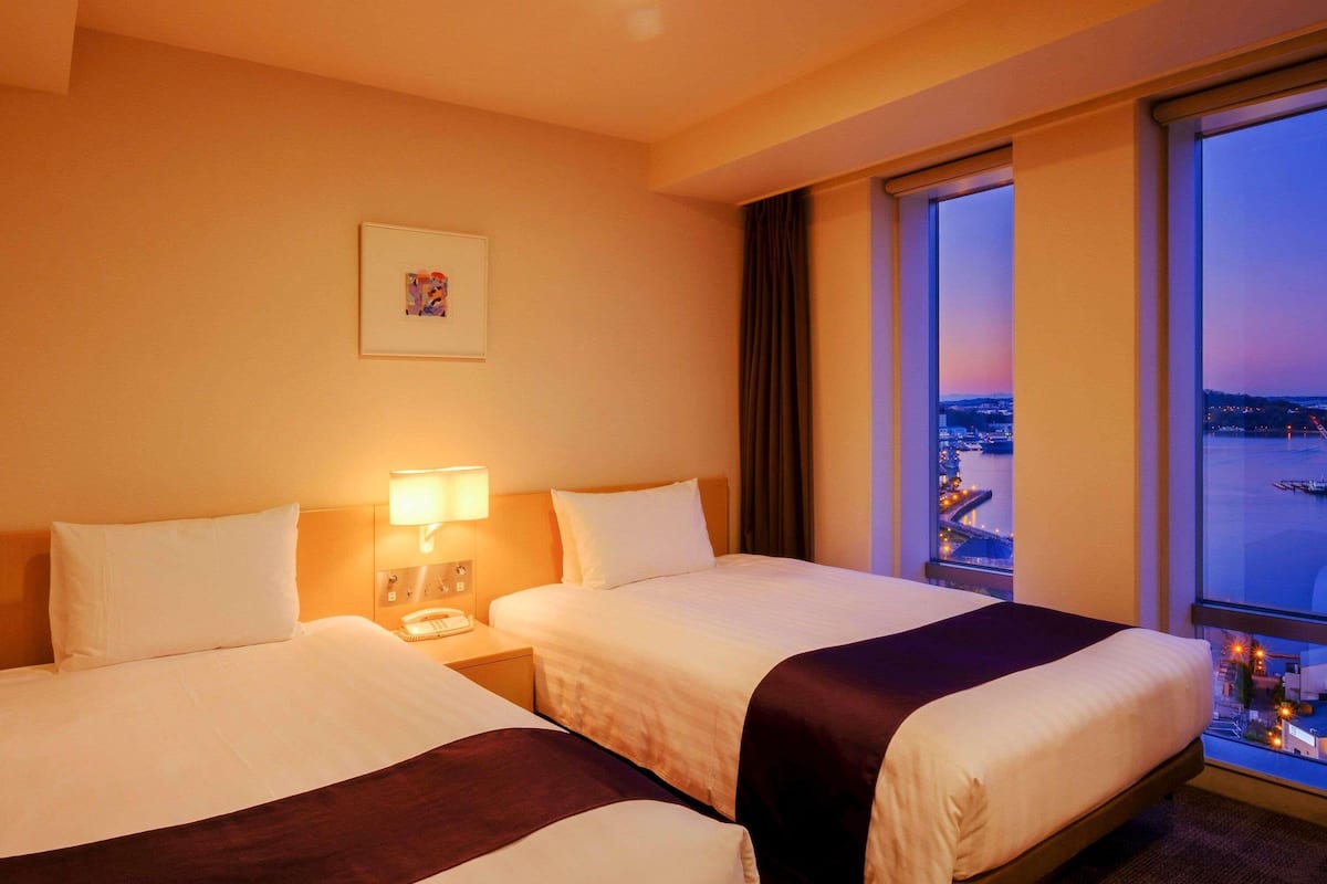 A hotel room with two beds, a nightstand with a lamp, a framed picture on the wall, and a large window offering a city view.