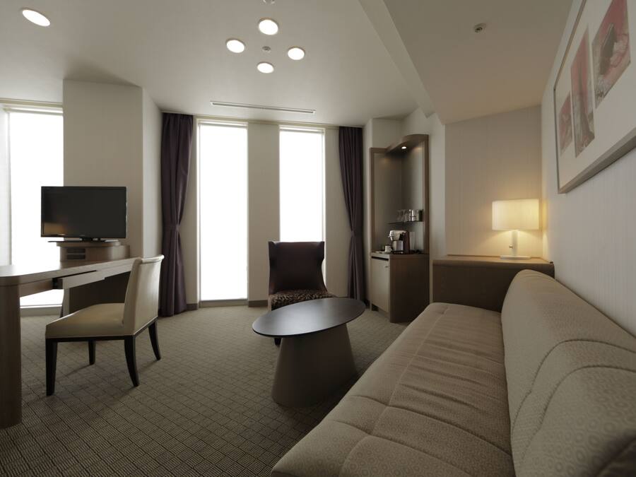 Habitación de hotel con sofá, escritorio y silla, televisión, lámpara y un amplio ventanal con cortinas.