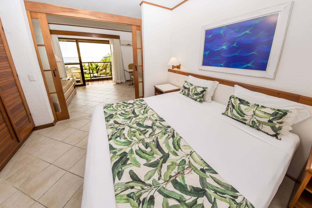 A hotel room with a bed, a nightstand, a lamp, a painting, and a view of the outdoors.