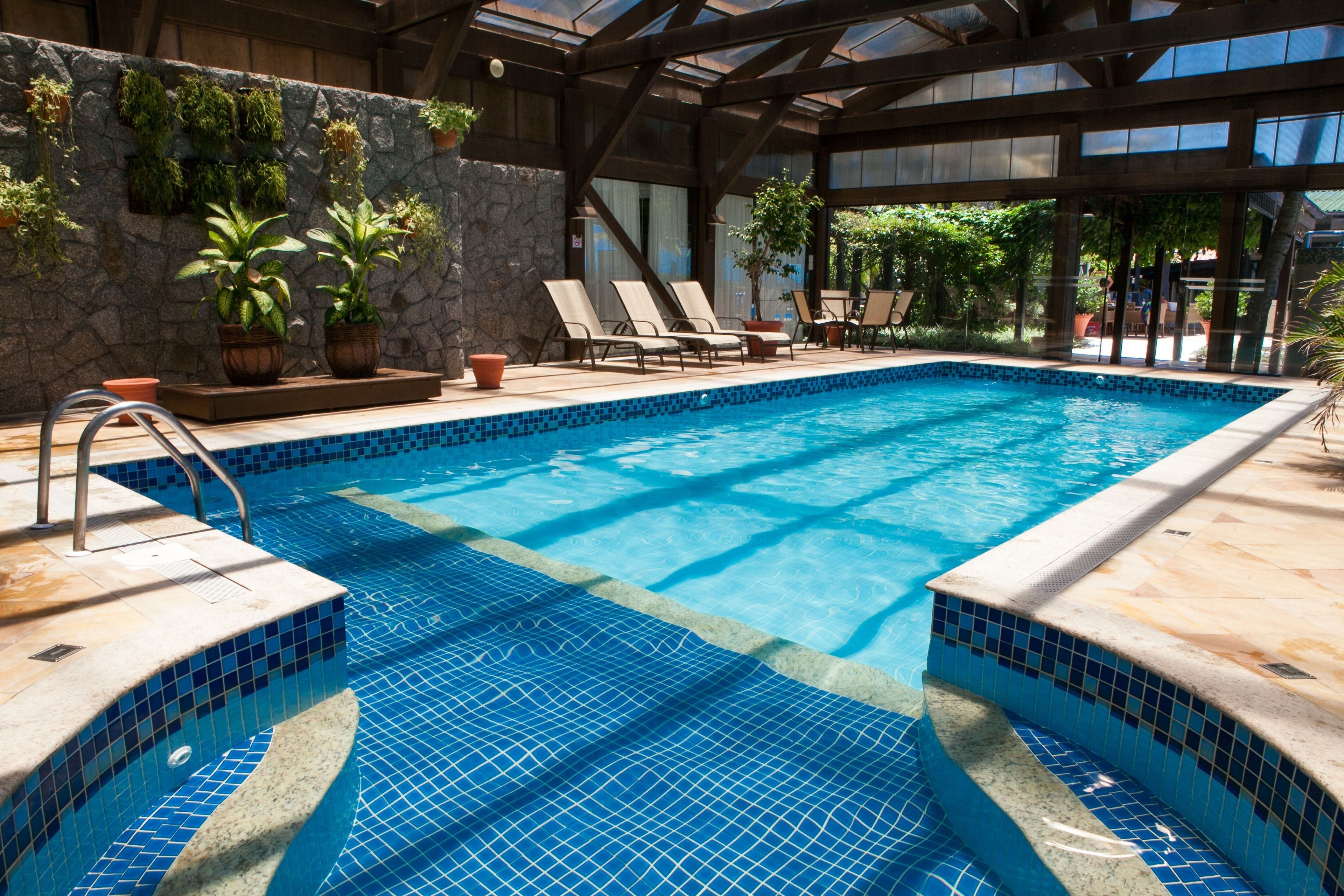 Una piscina interior con bordes de azulejos azules, sillas de descanso y plantas en macetas.