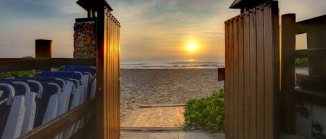 Una entrada frente a la playa con un portón de madera que da a una zona de arena, un muro de piedra y una fila de sillas azules.