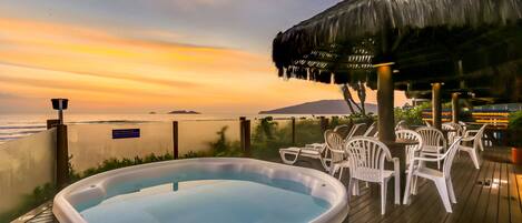 A hot tub on a wooden deck with white plastic chairs, overlooking a body of water and a sunset.