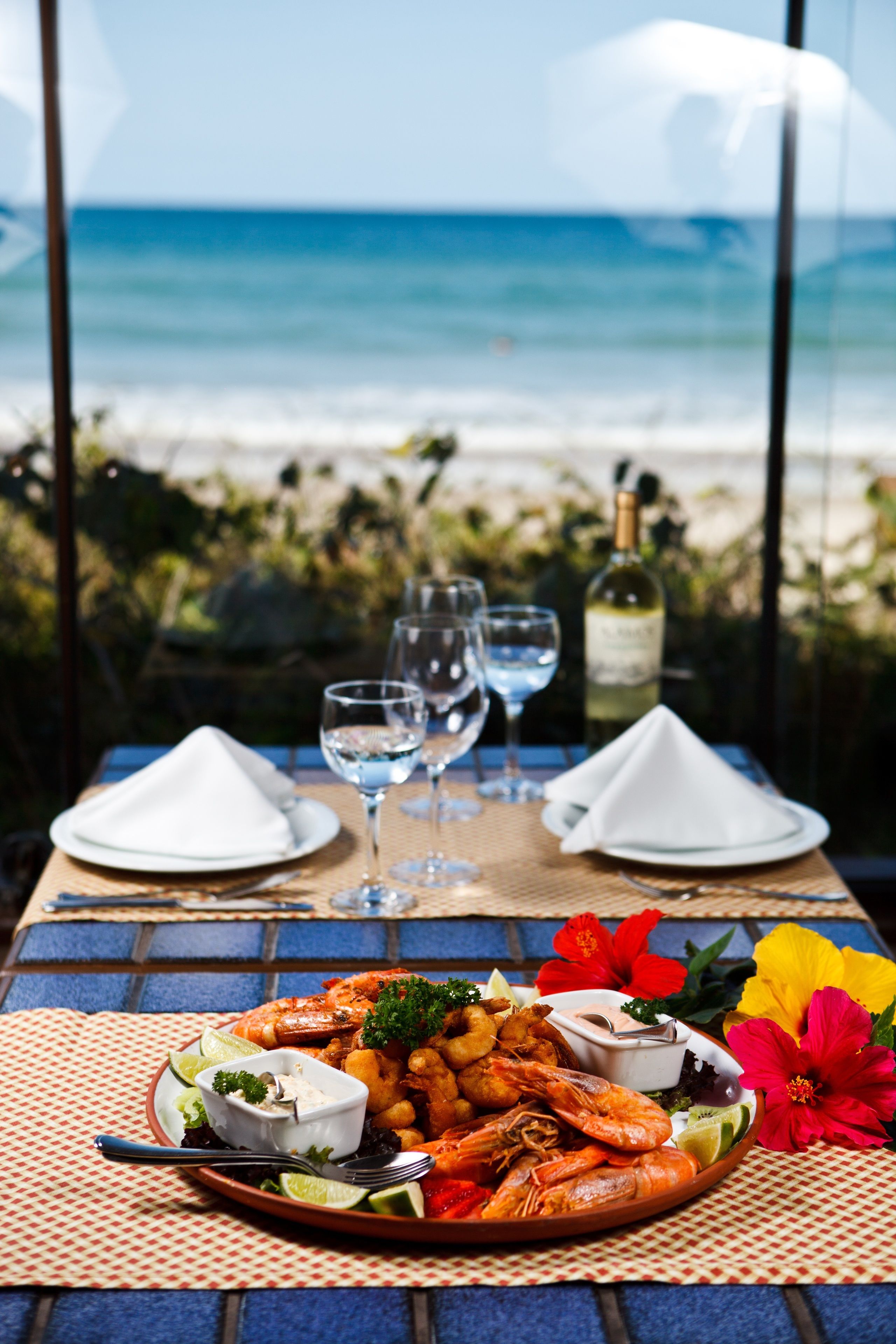 Mesa com frutos do mar, copos e uma garrafa, com vista para a praia.