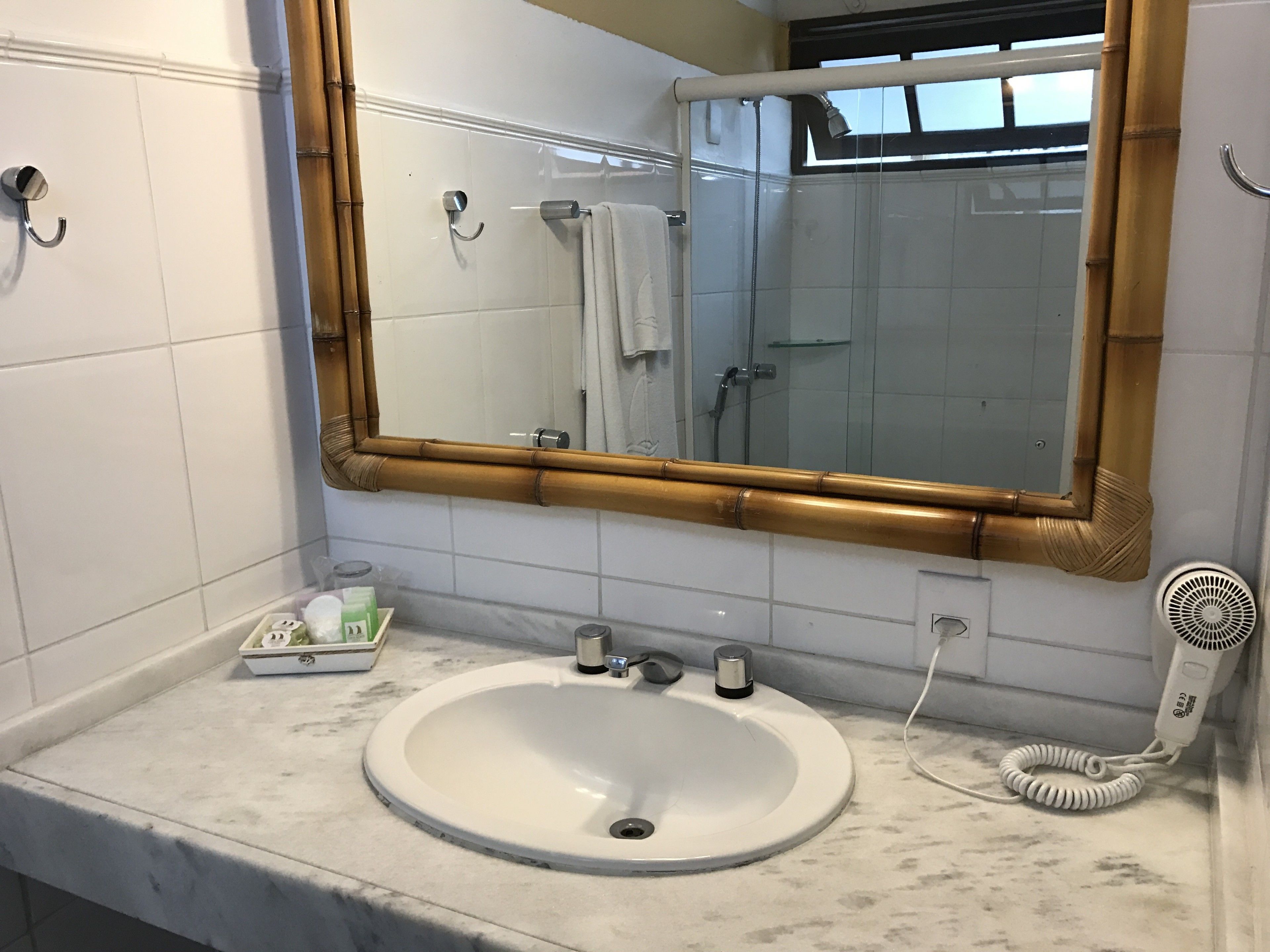 Un baño con un lavamanos blanco, un espejo con marco de bambú, un perchero y un secador de pelo en la encimera.