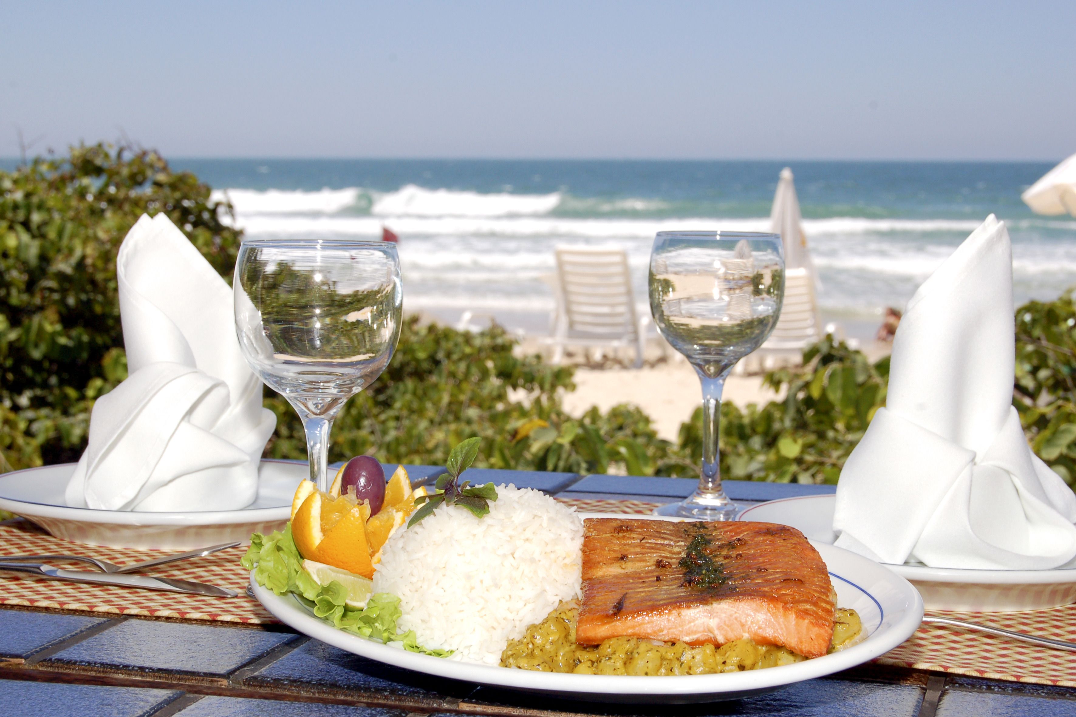 Une table dressée avec un repas composé de saumon, de riz et de légumes, deux verres à vin, et une vue sur l’océan.