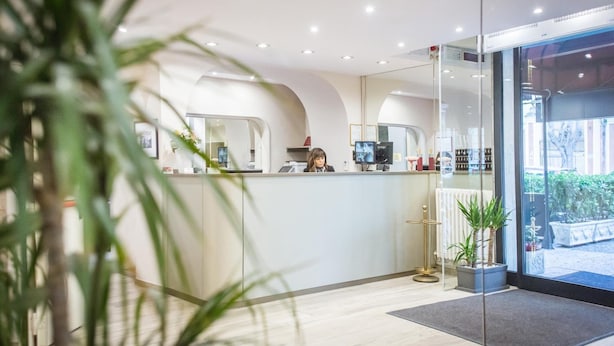 A reception area with a receptionist, a glass partition, a potted plant, and a view of the outdoor area. - Europa (Modena)
