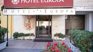 The entrance of Hotel Europa with a red awning, a glass door, and a flower bed with red flowers. - Europa (Modena)