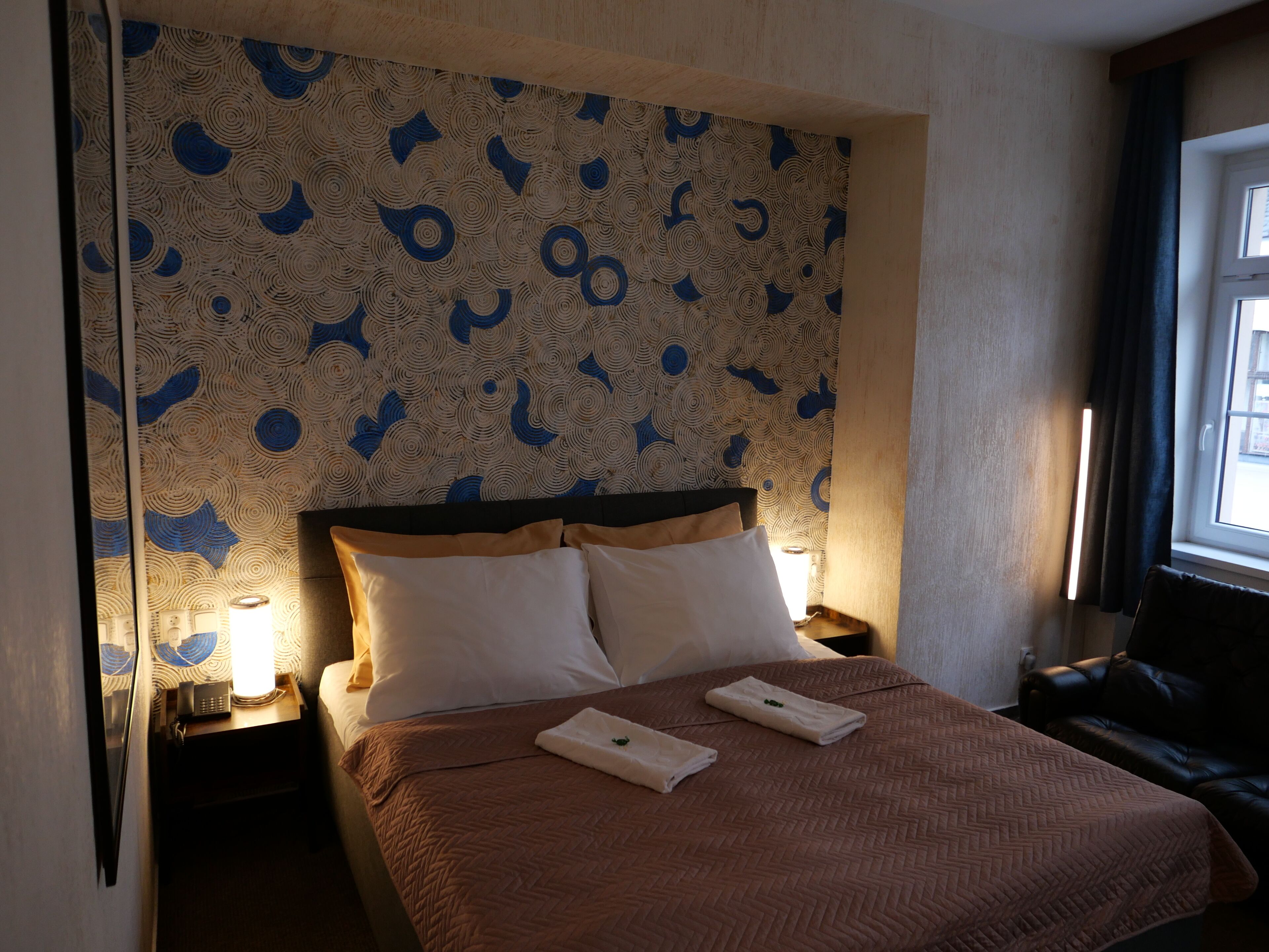 A hotel room with a patterned wallpaper, a bed with two pillows, and a nightstand with a lamp.