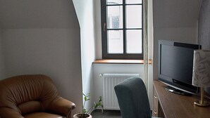 A small, well-lit room with a brown leather chair, a blue armchair, a wooden desk with a television, and a window with a view of the city. - Hotel Radnice (Liberec)