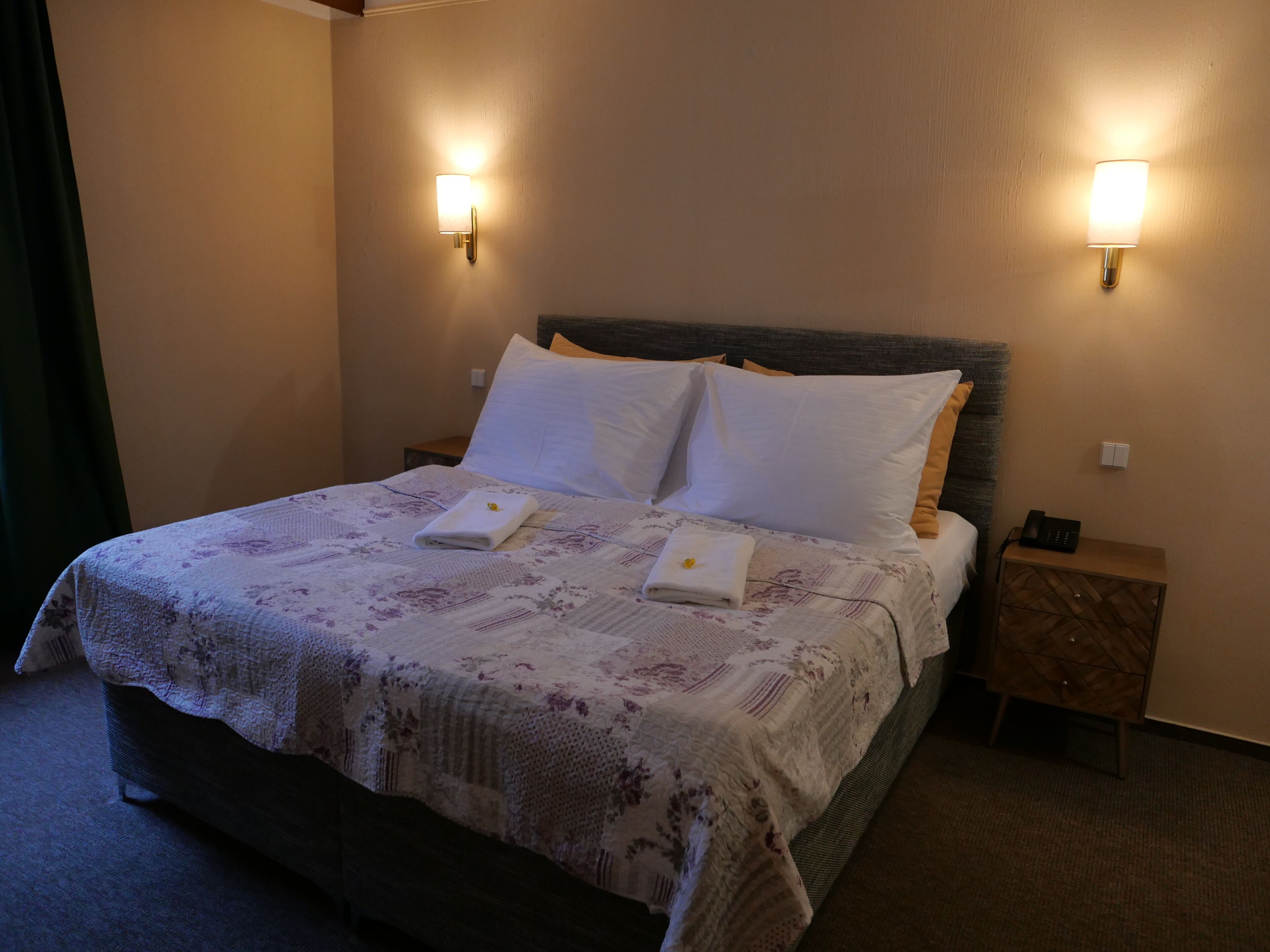 A neatly made bed with a floral bedspread, two pillows, and two folded towels. There are two wall-mounted light fixtures on either side of the bed.