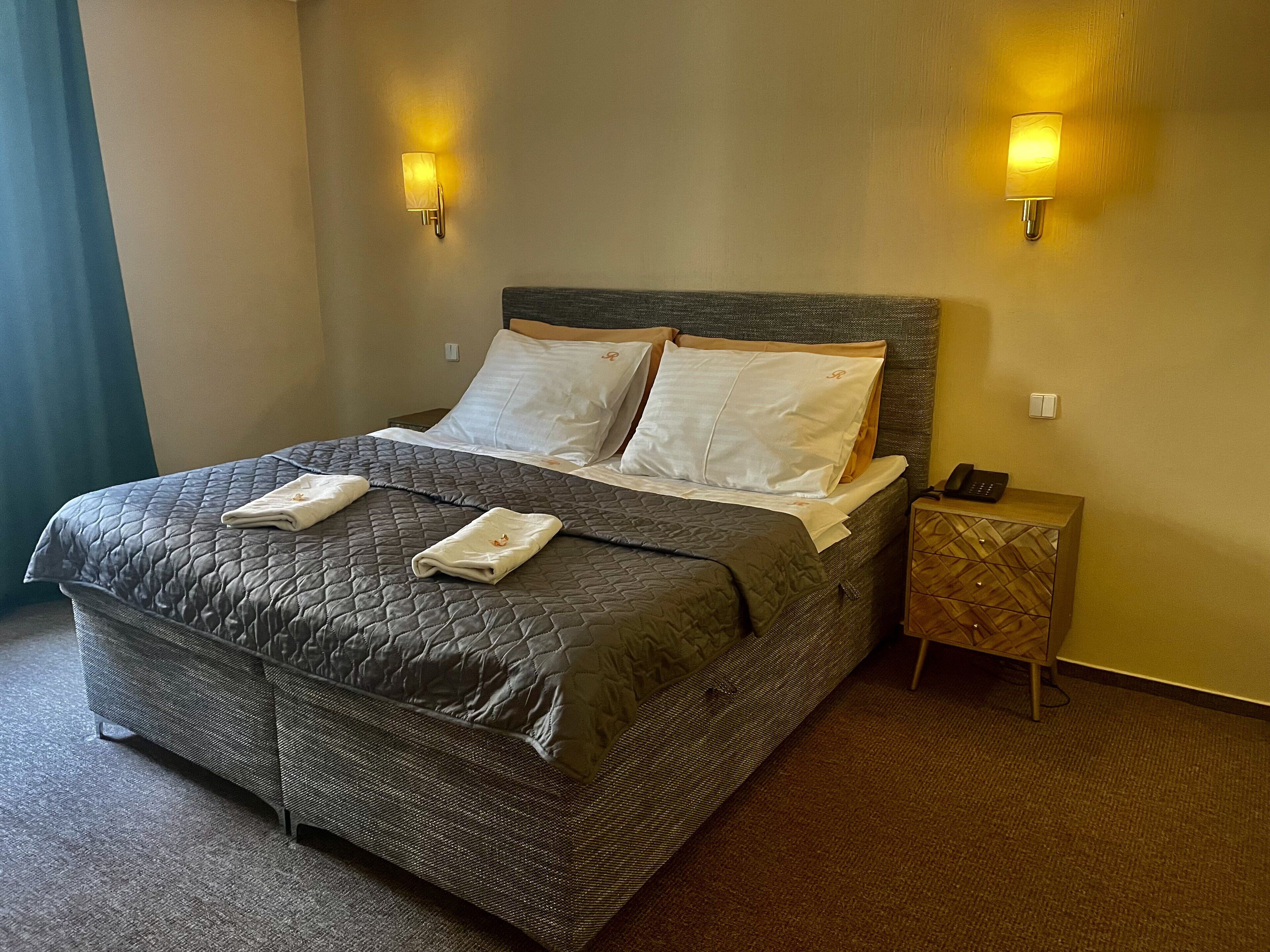 A neatly made bed with a quilted headboard, two bedside lamps, a bedside phone, and a small wooden nightstand with a patterned drawer.