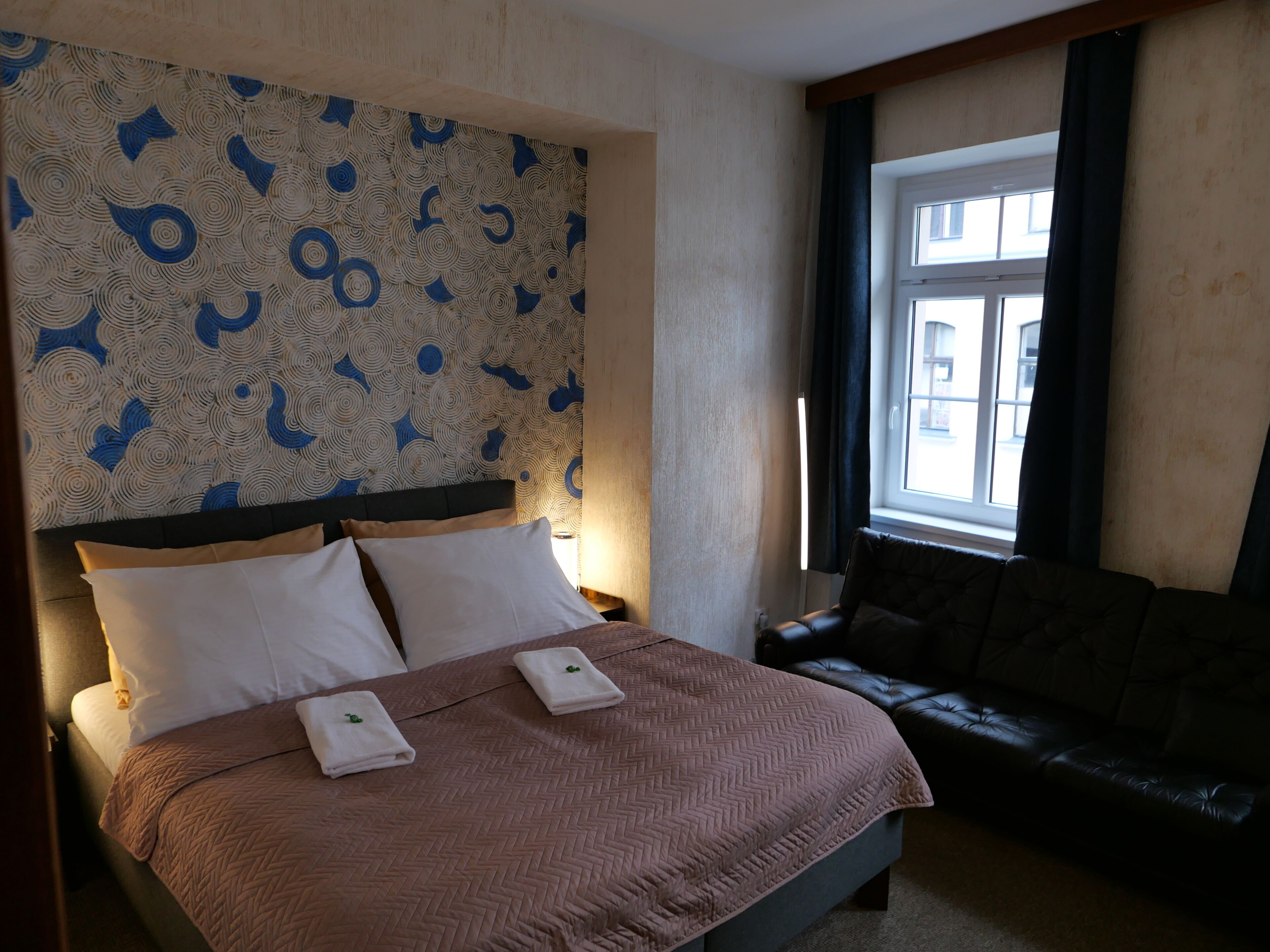 Habitación de hotel con cama, cabecero con papel tapiz estampado, ventana con cortinas y sofá de cuero oscuro.