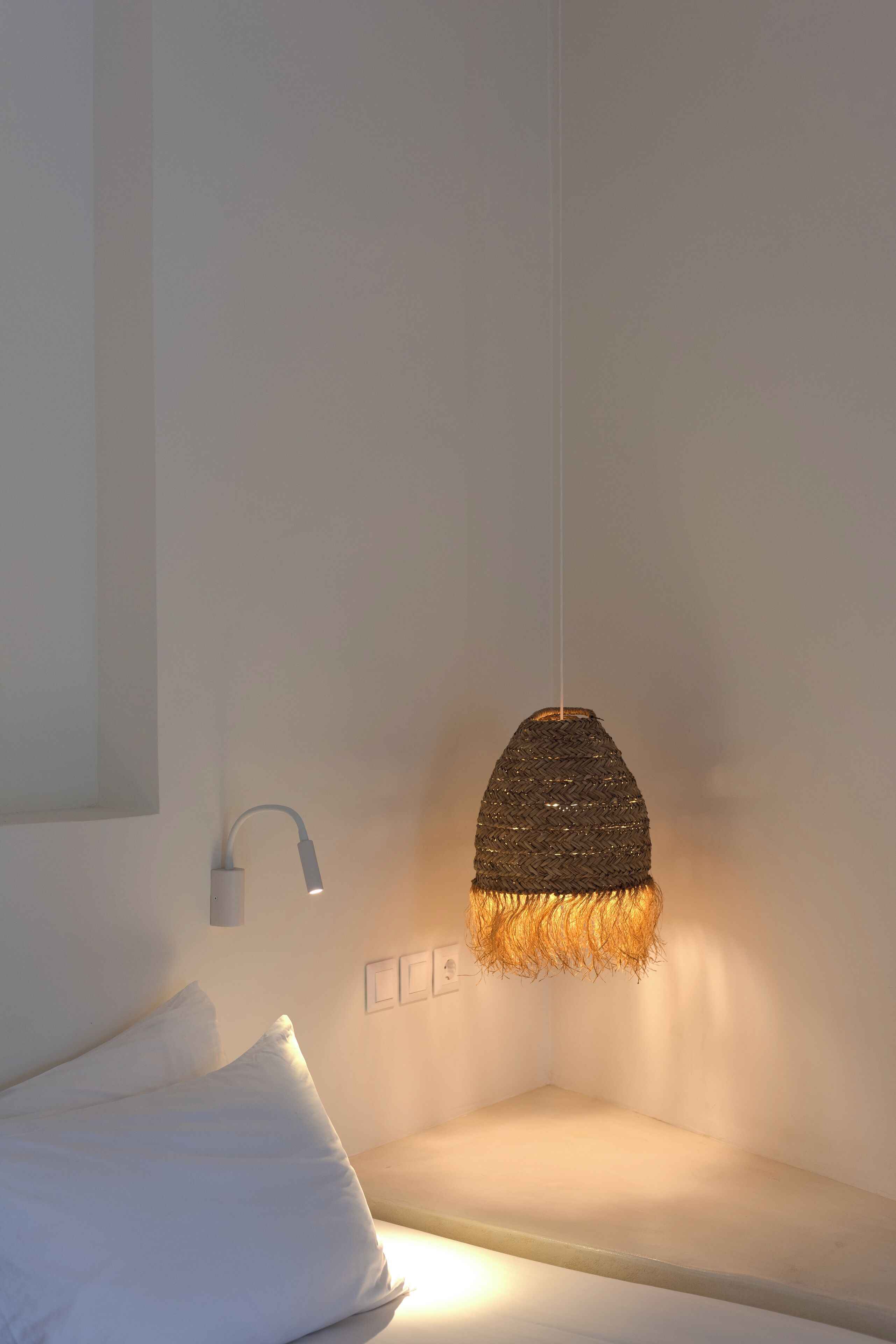 A minimalist hotel room with a bed, a wall-mounted lamp, and a textured wall hanging.