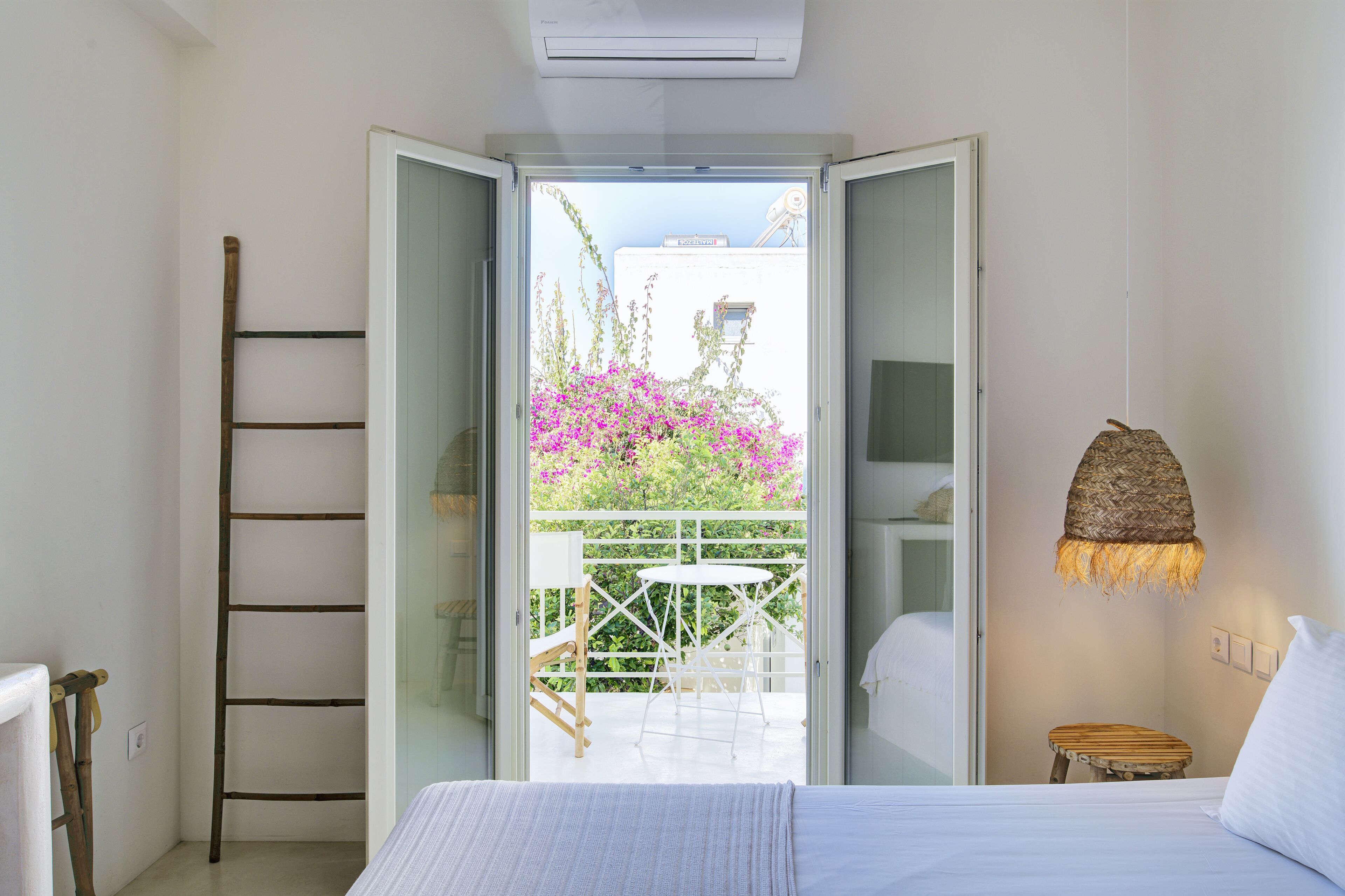 A bedroom with a bed, a wooden ladder, a hanging light fixture, and an open door leading to a balcony with a table and chairs.