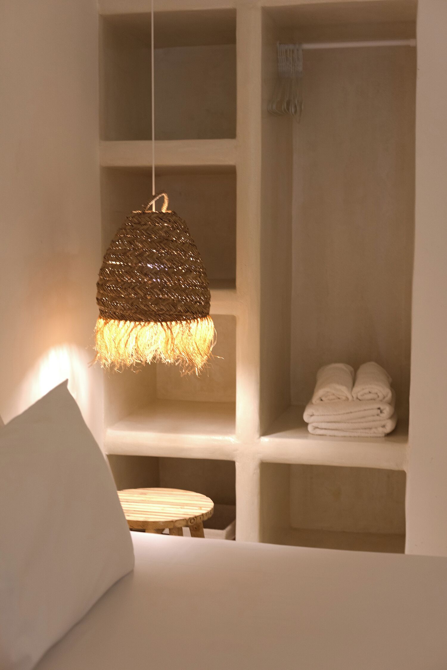 Une pièce avec un luminaire suspendu, un lit, un petit tabouret en bois et une étagère où sont pliées des serviettes.
