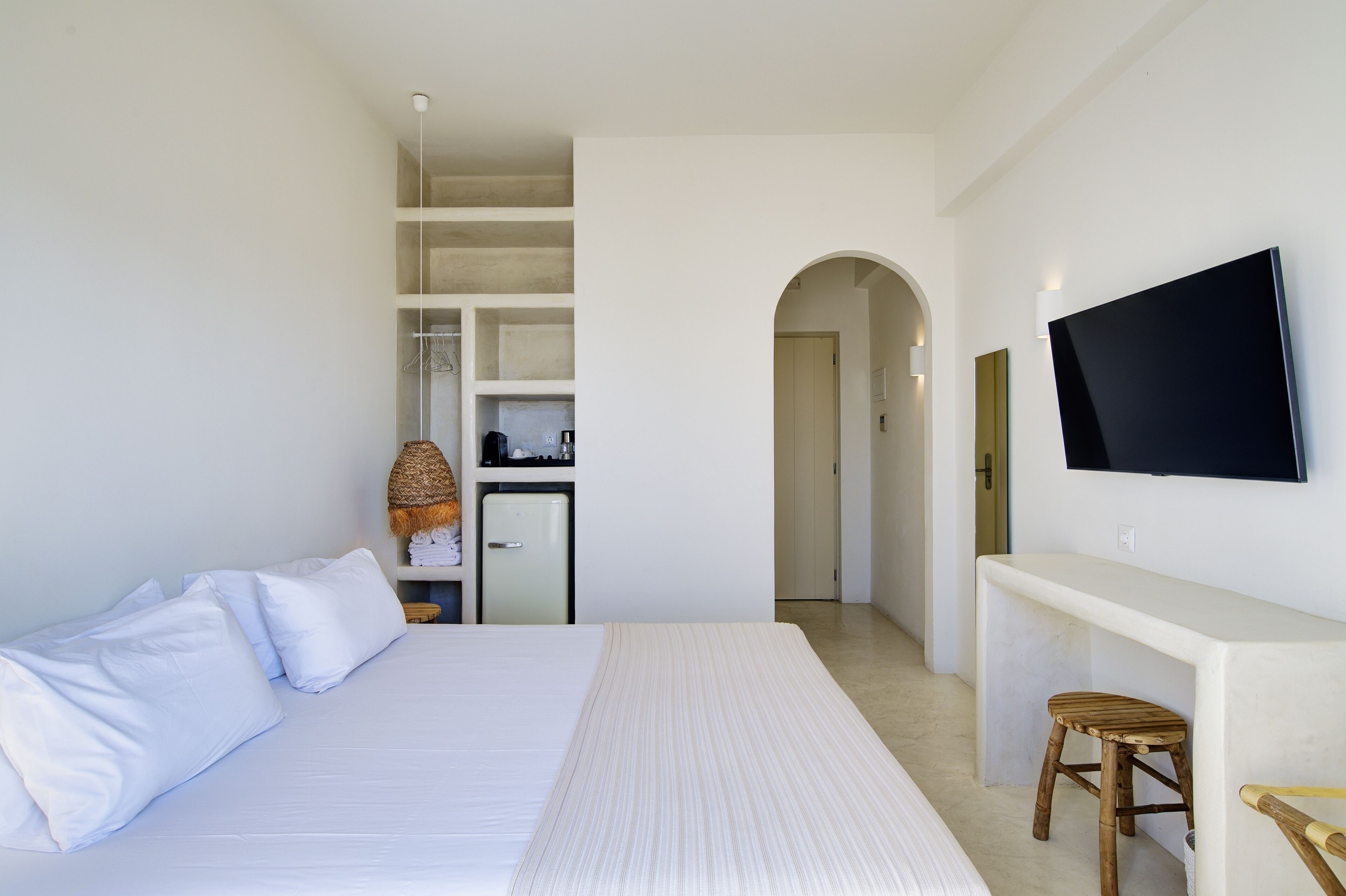 A hotel room with a large bed, a TV mounted on the wall, a small kitchenette, and a wooden stool.