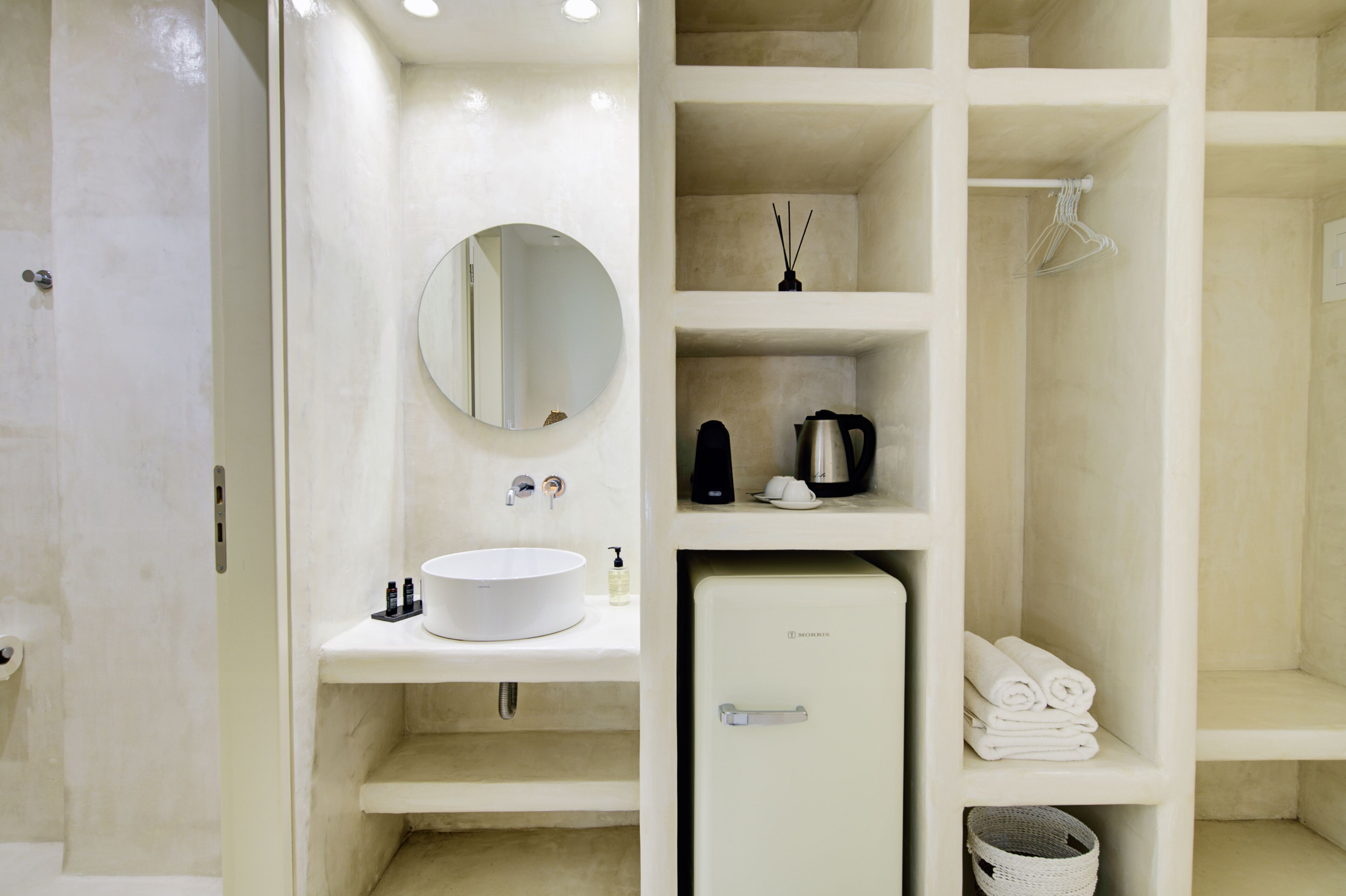 A modern bathroom with a white sink, mirror, and shelves containing toiletries and towels.
