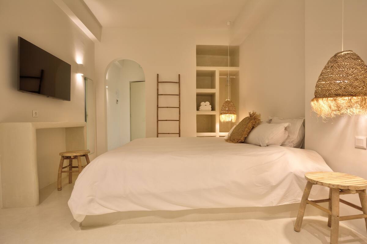 A bedroom with a large bed, a television mounted on the wall, a wooden ladder, and a woven light fixture.